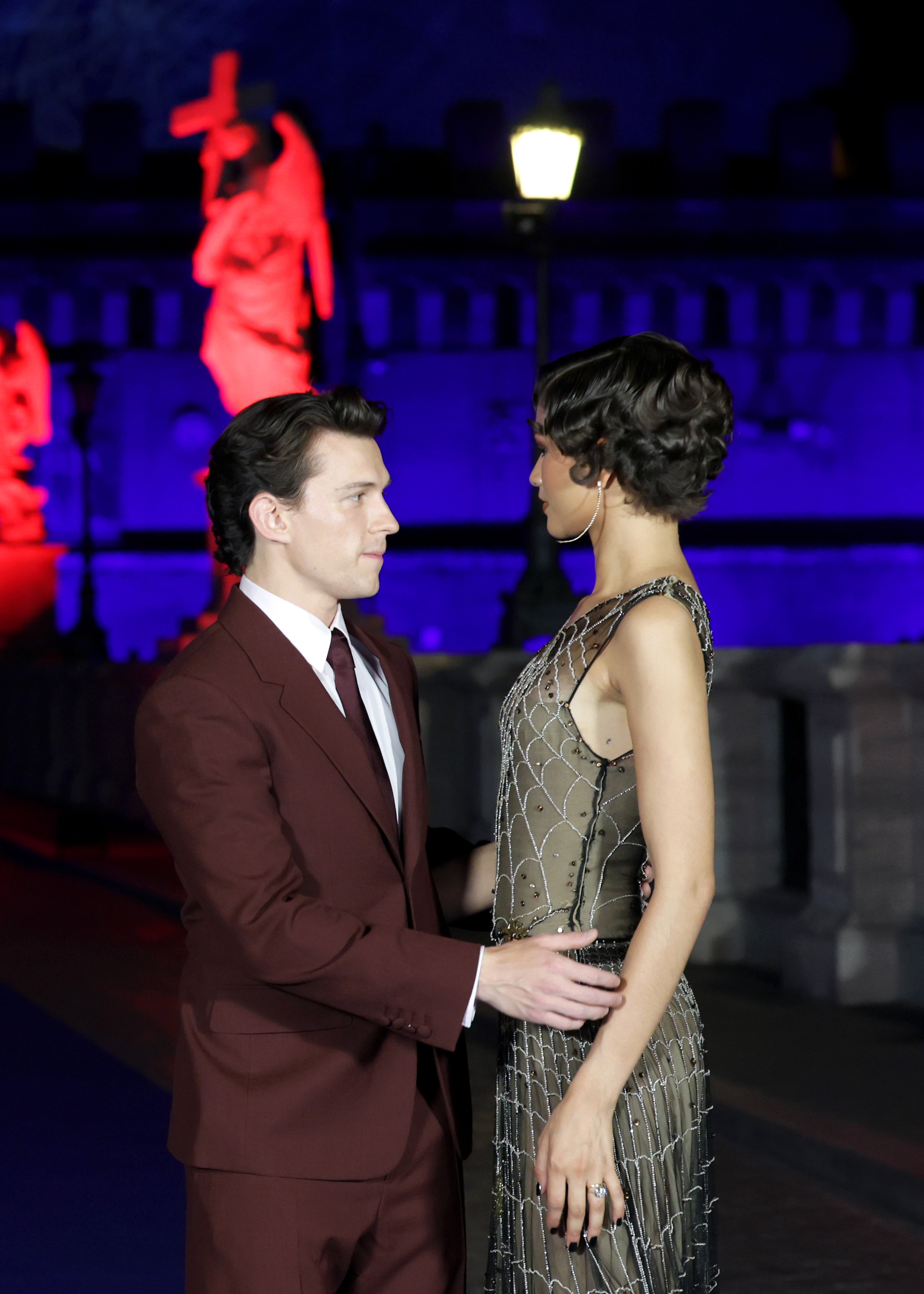 Two individuals in elegant attire, one in a tailored suit and the other in a beaded gown, share a moment at a nighttime event