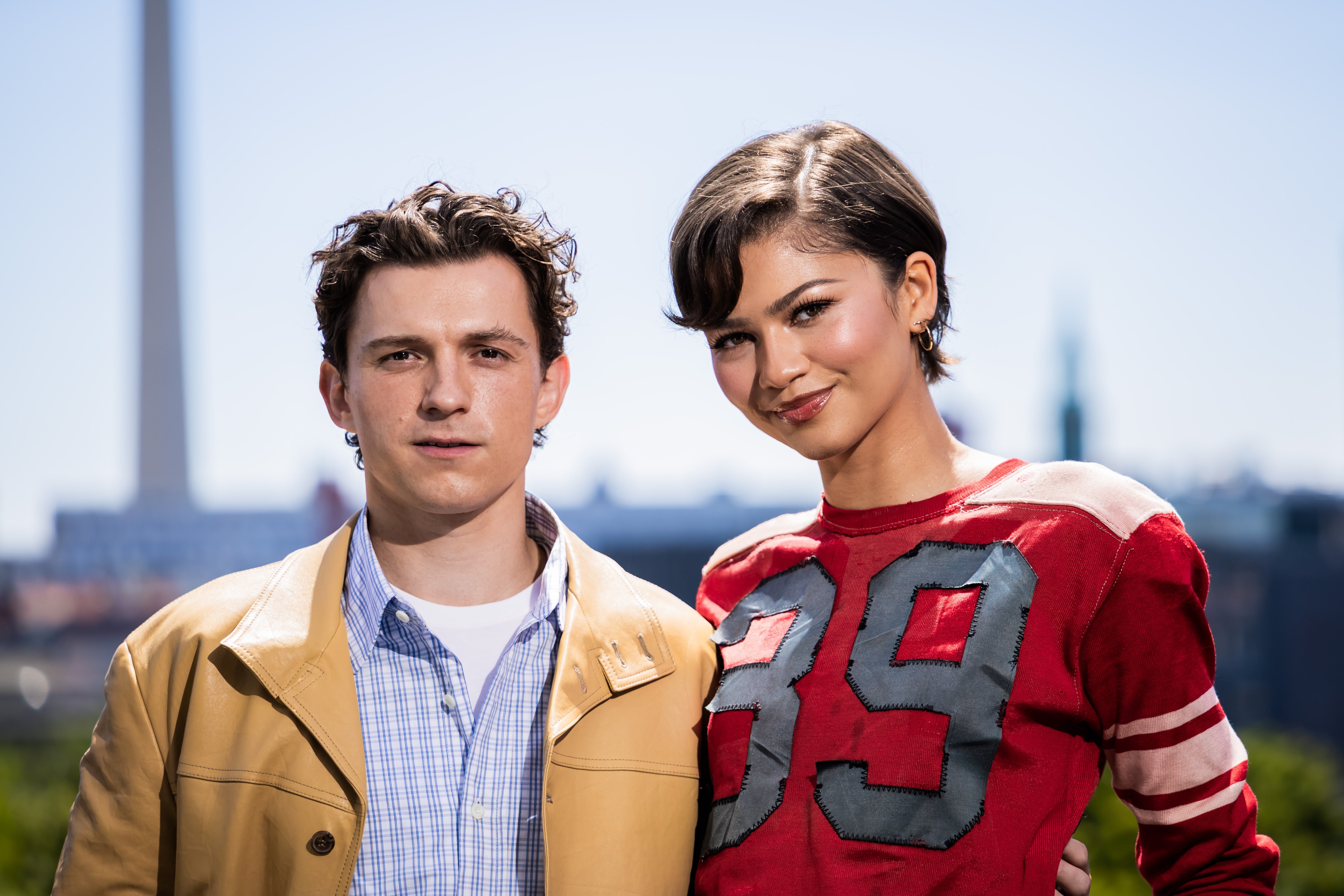 Two individuals outdoors, 1 successful a casual overgarment and shirt, the different successful a apical with "39" connected it, posing unneurotic with a cityscape successful the background