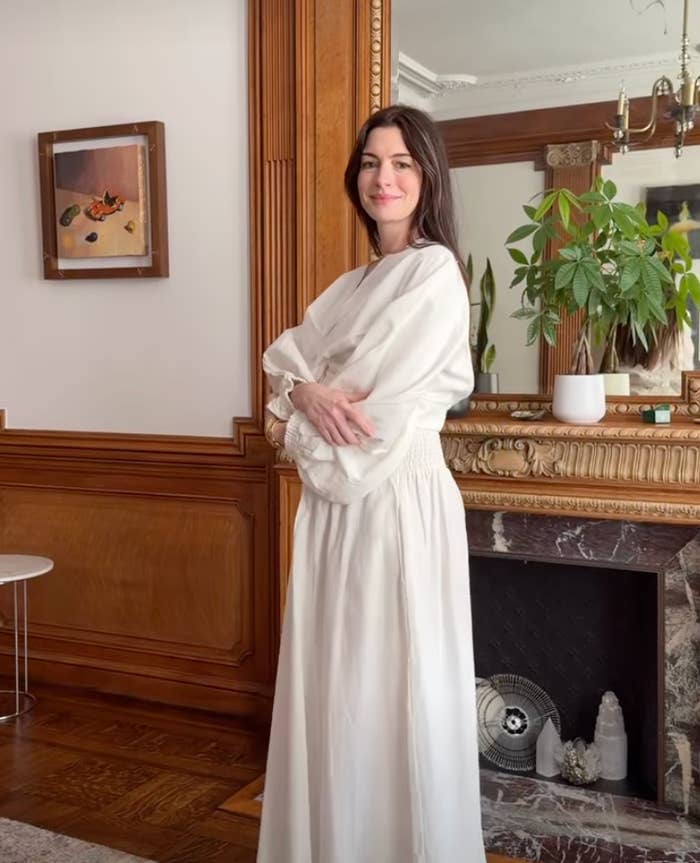 Anne Hathaway in a long, flowing dress stands in a stylish room with a plant and a decorated mantel