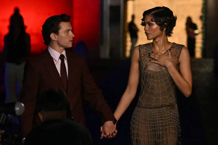 A mates  connected  a reddish  carpet, holding hands. One wears a suit; the different   successful  a geometric-patterned, sheer dress