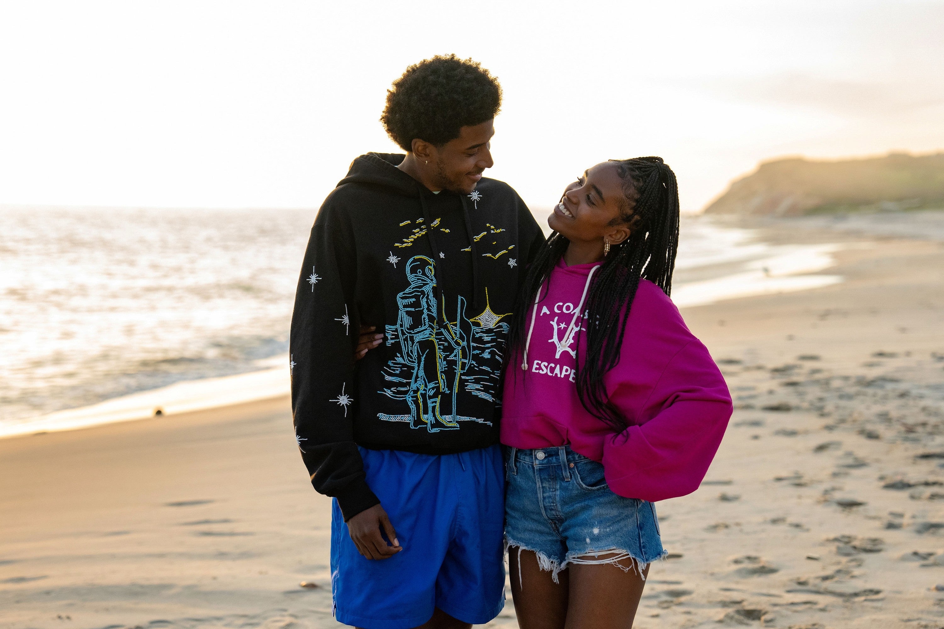 Two radical   smiling astatine  each   different   connected  a beach; 1  wearing an astronaut-themed hoodie, the different   successful  a pinkish  sweatshirt and denim shorts