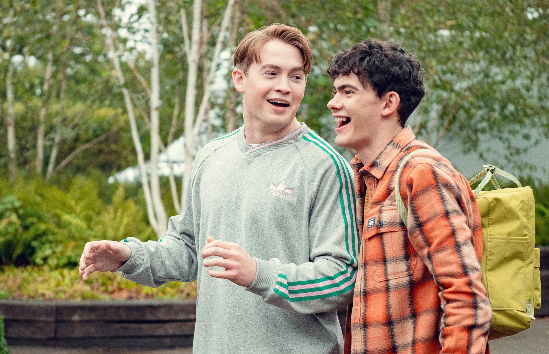 Two radical   walking outdoors, smiling and talking. One wears a casual tracksuit, the different   wears a plaid garment  and backpack