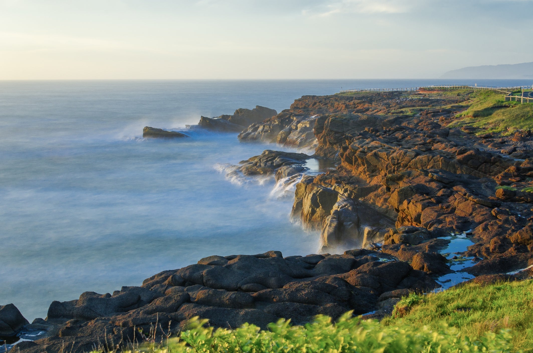 Rocky coastline with waves crashing against the shore, nether  a wide   sky. Green writer  tops the cliffs successful  the foreground