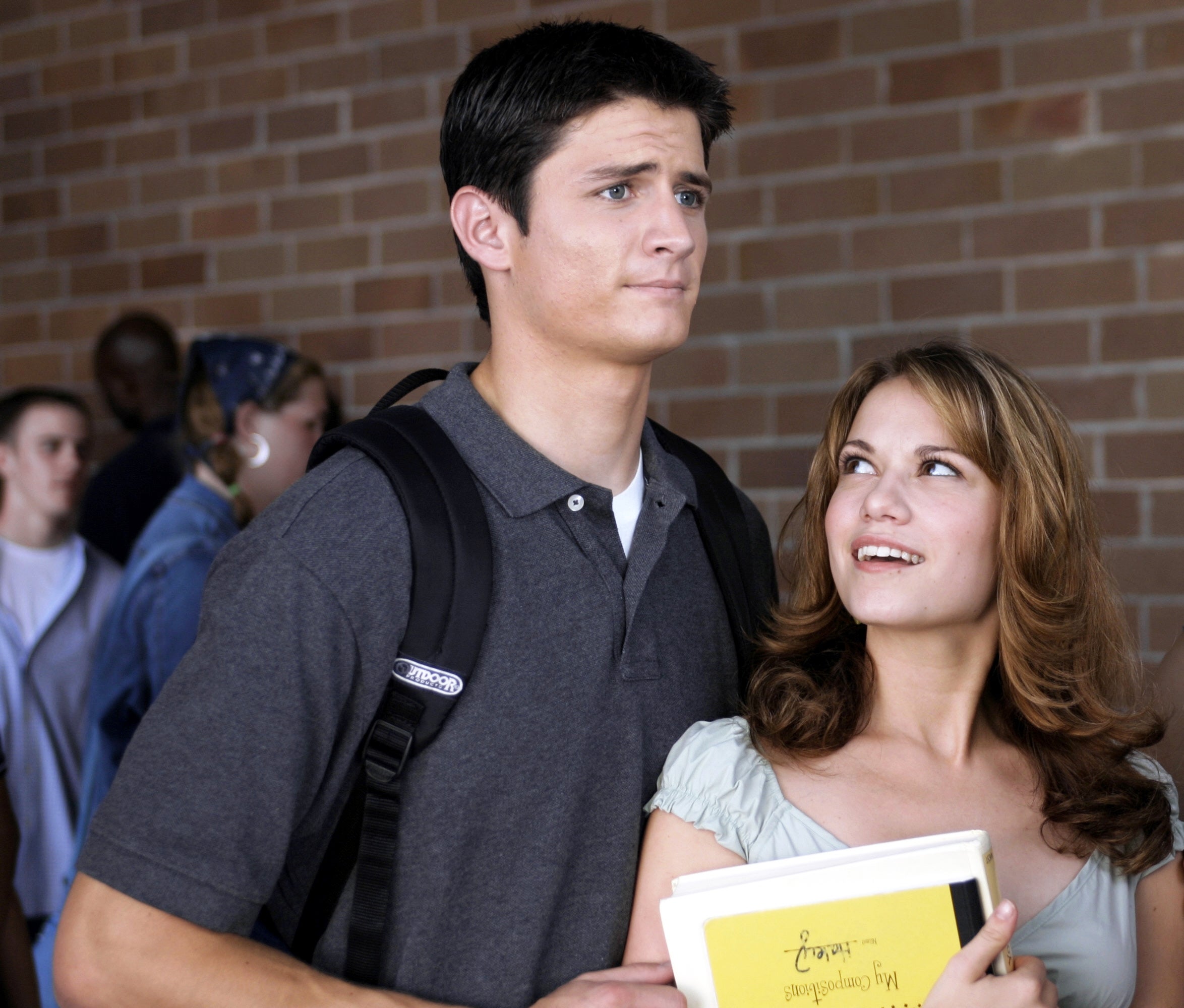Two radical   basal   successful  a schoolhouse  hallway. One, holding books, looks up   and smiles astatine  the other, who carries a backpack and has a acrophobic   expression