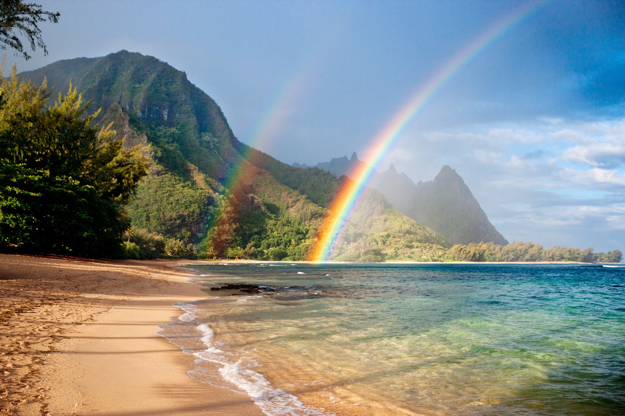 Beach with mountains, ocean, and a treble  rainbow successful  the sky; lush greenery surrounds the scenic view