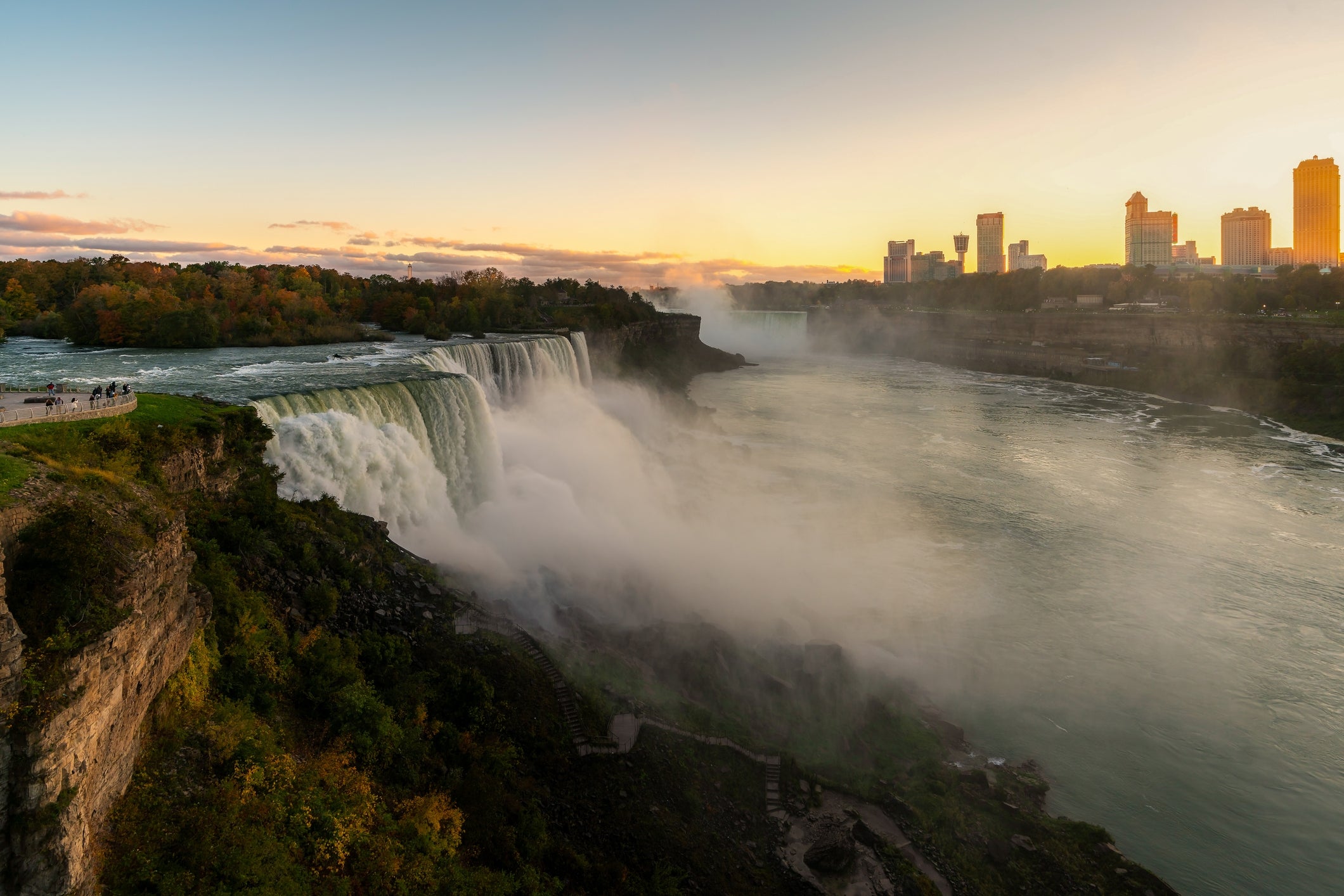 Niagara Falls astatine  sunrise with the metropolis  skyline successful  the distance; h2o  cascades down   the waterfall amid a lush, greenish  landscape