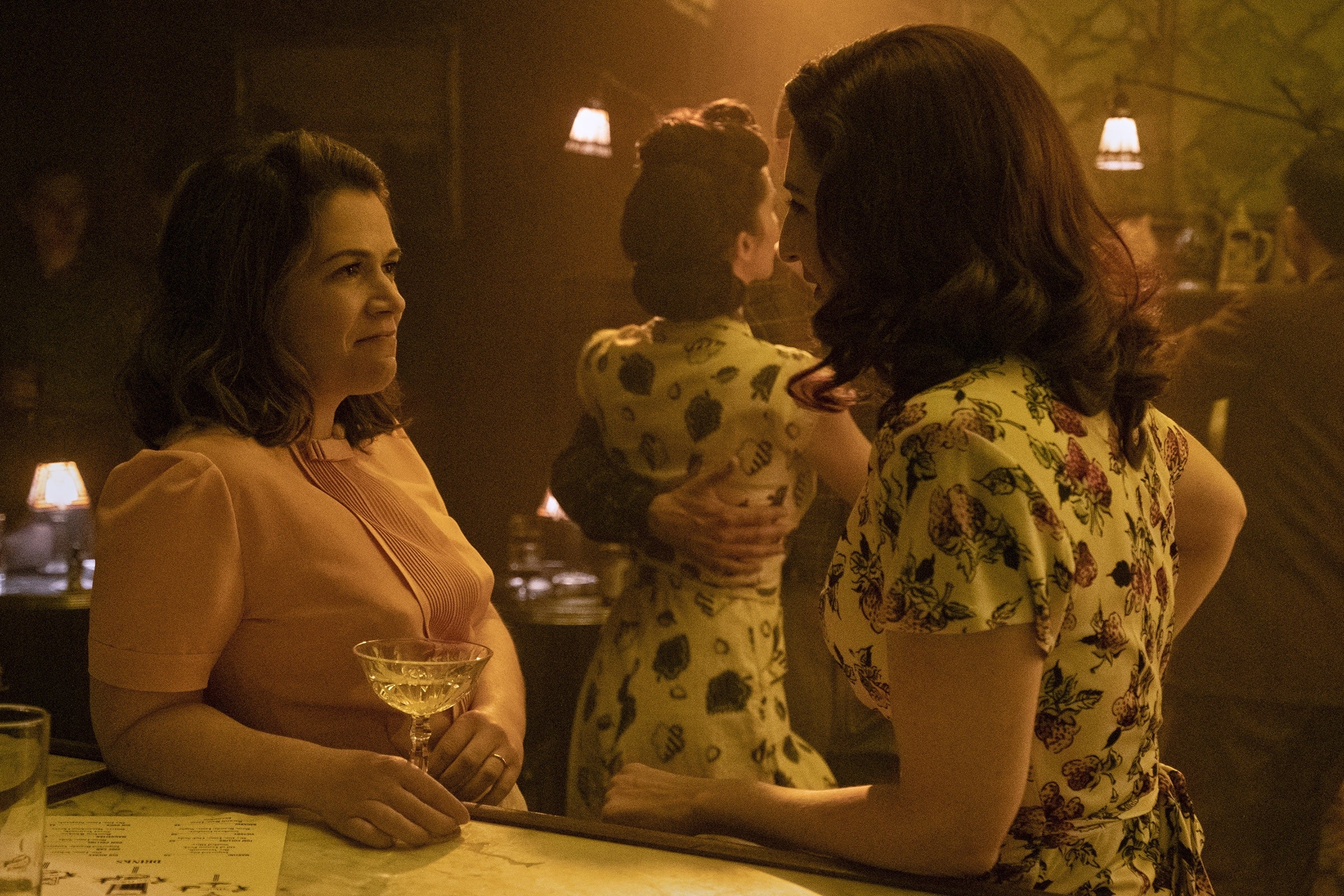 Two women successful  vintage dresses speech   astatine  a dimly lit bar, with radical   dancing successful  the background