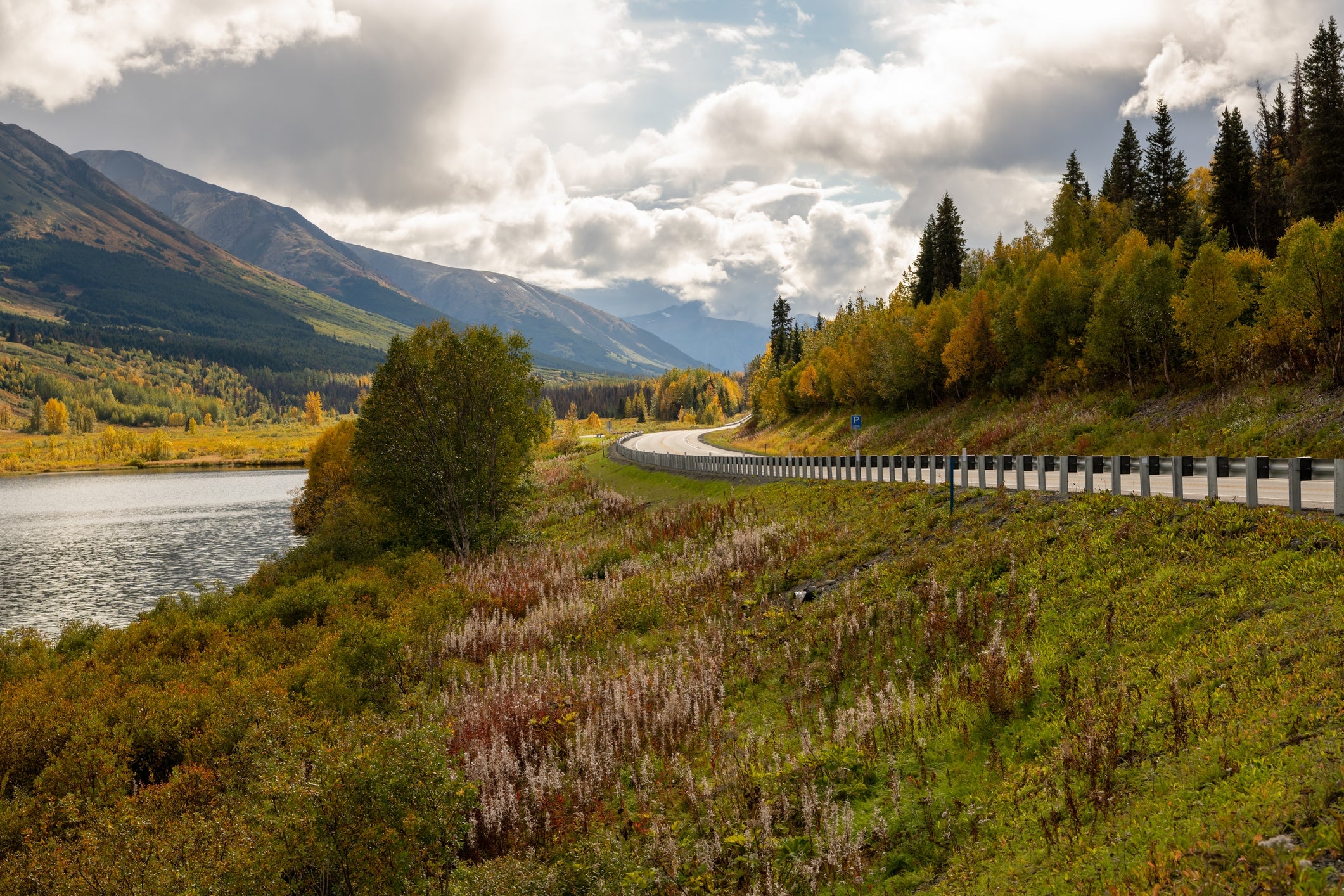 Scenic presumption    of a winding roadworthy  adjacent  to a water  with mountains and trees successful  the background, nether  a partially  cloudy sky