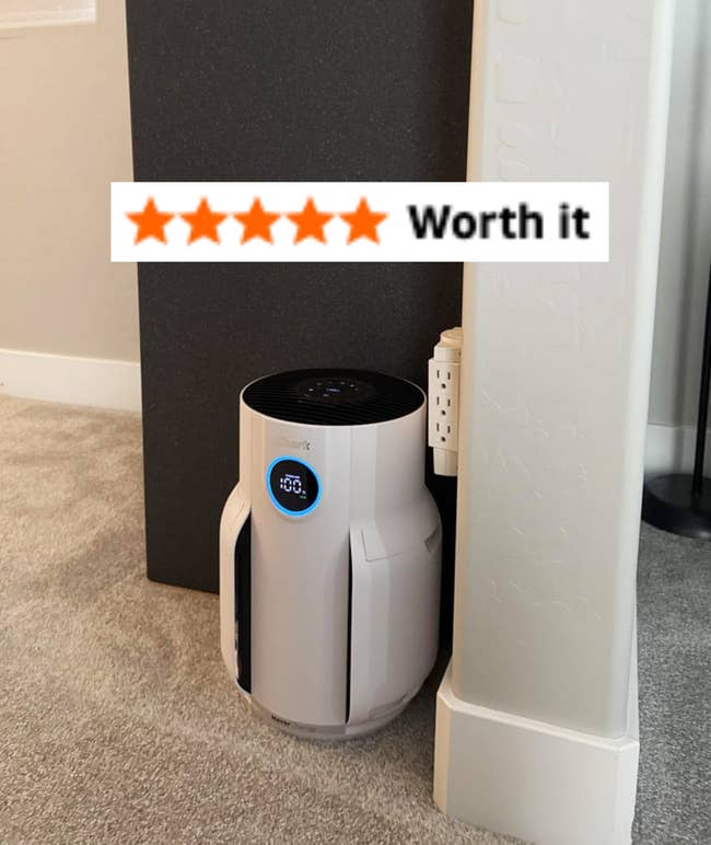 Compact, modern air purifier positioned in a home corner, plugged into an outlet, with a digital display showing 100% air quality