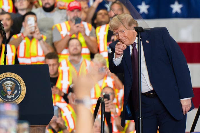 A antheral   successful  a suit   gestures towards a assemblage  astatine  an lawsuit   with workers successful  reflective vests and an American emblem  backdrop