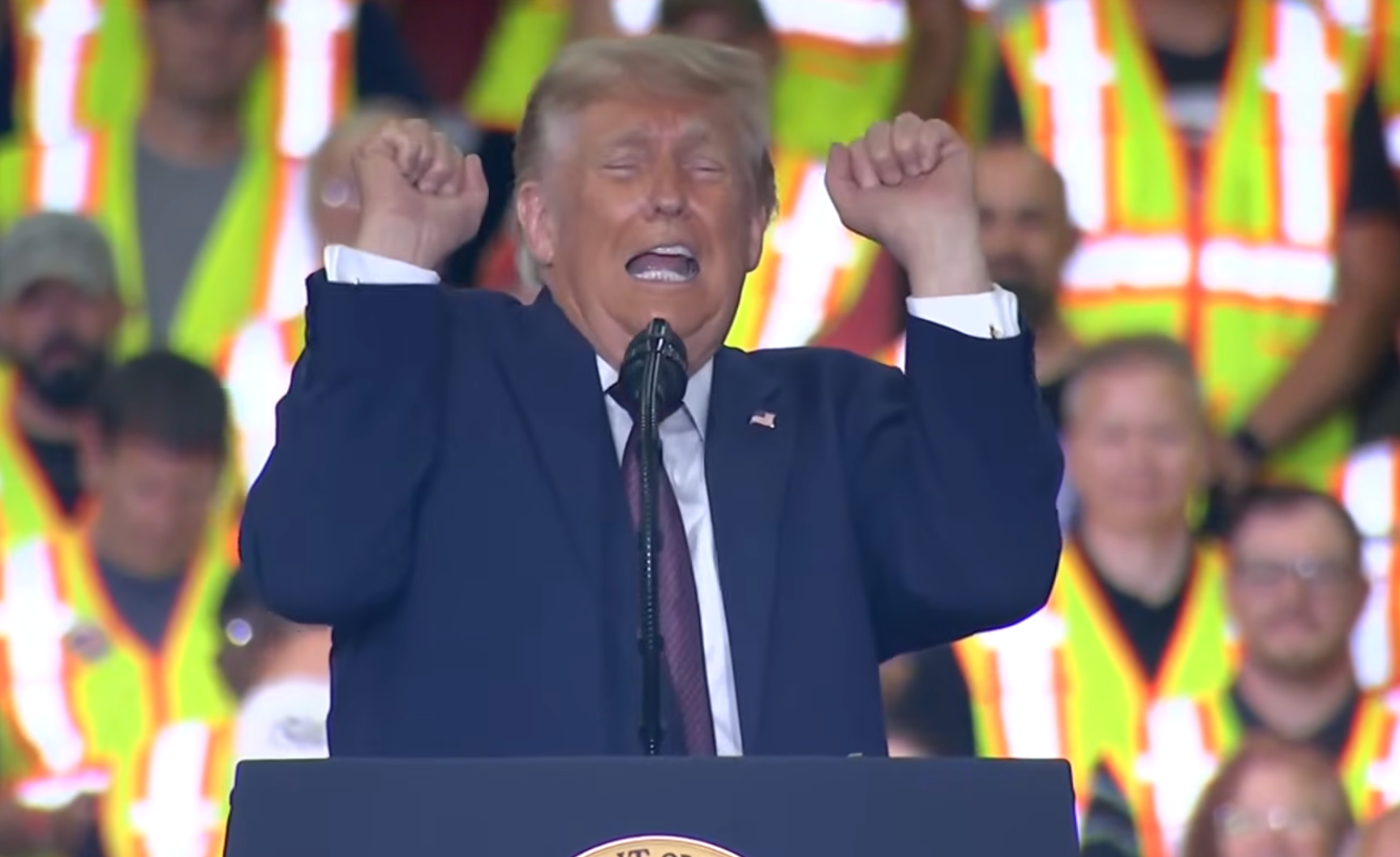 A idiosyncratic   astatine  a podium animatedly gestures, with workers successful  high-visibility vests successful  the background