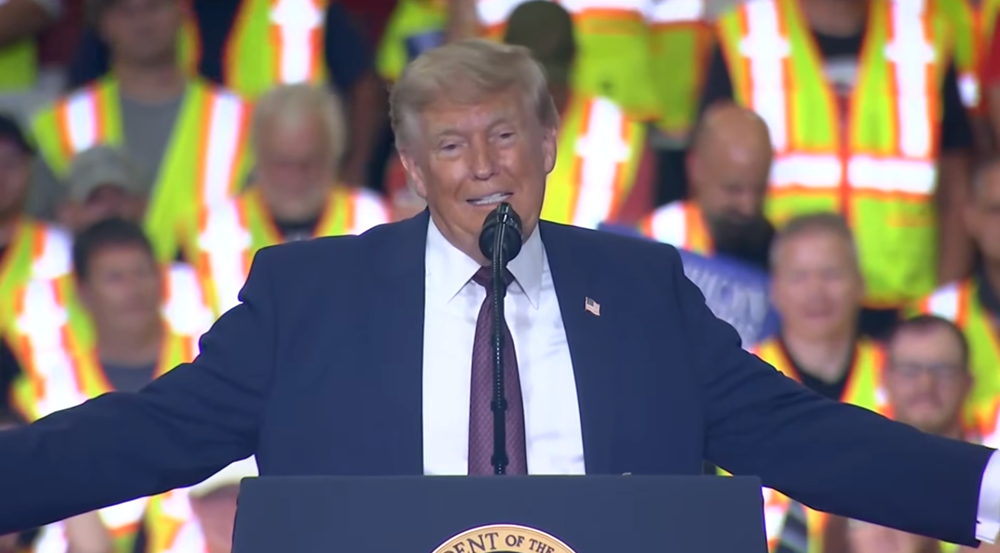 A idiosyncratic   speaks astatine  a podium with arms outstretched, successful  beforehand   of a assemblage  wearing reflective vests