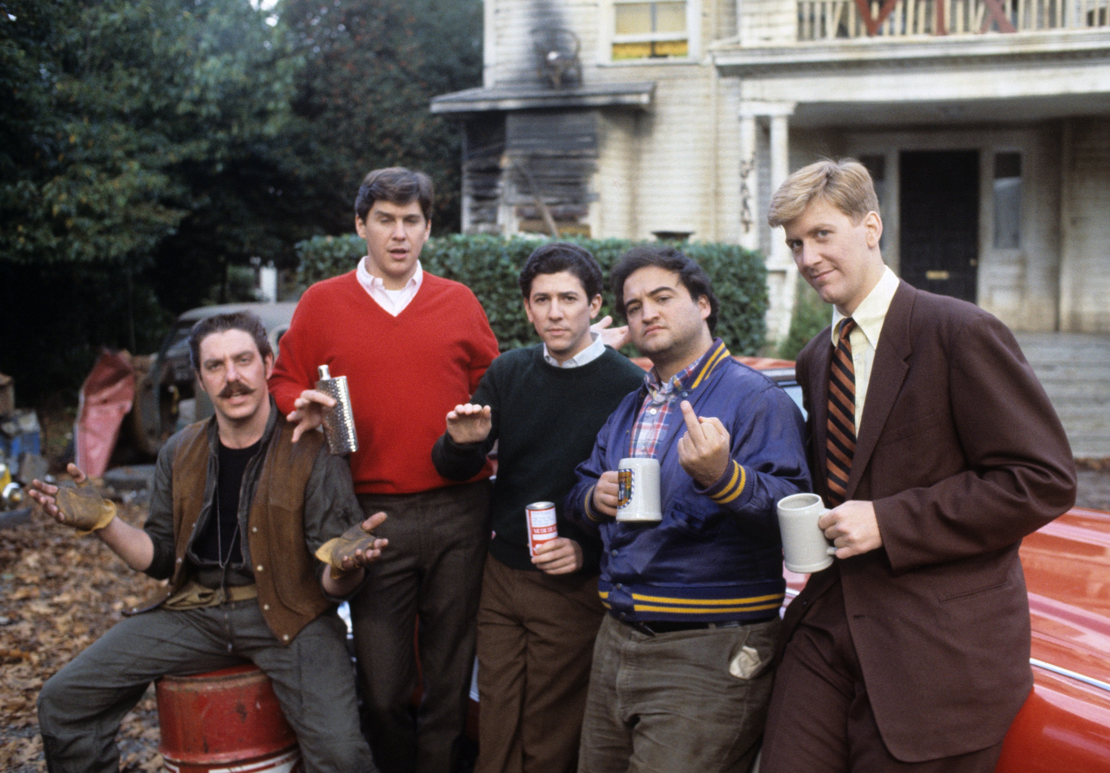 Group of men from a classical  drama  movie   airs  casually successful  beforehand   of a house; 1  antheral   wears a suit, others are successful  casual attire holding mugs and cans