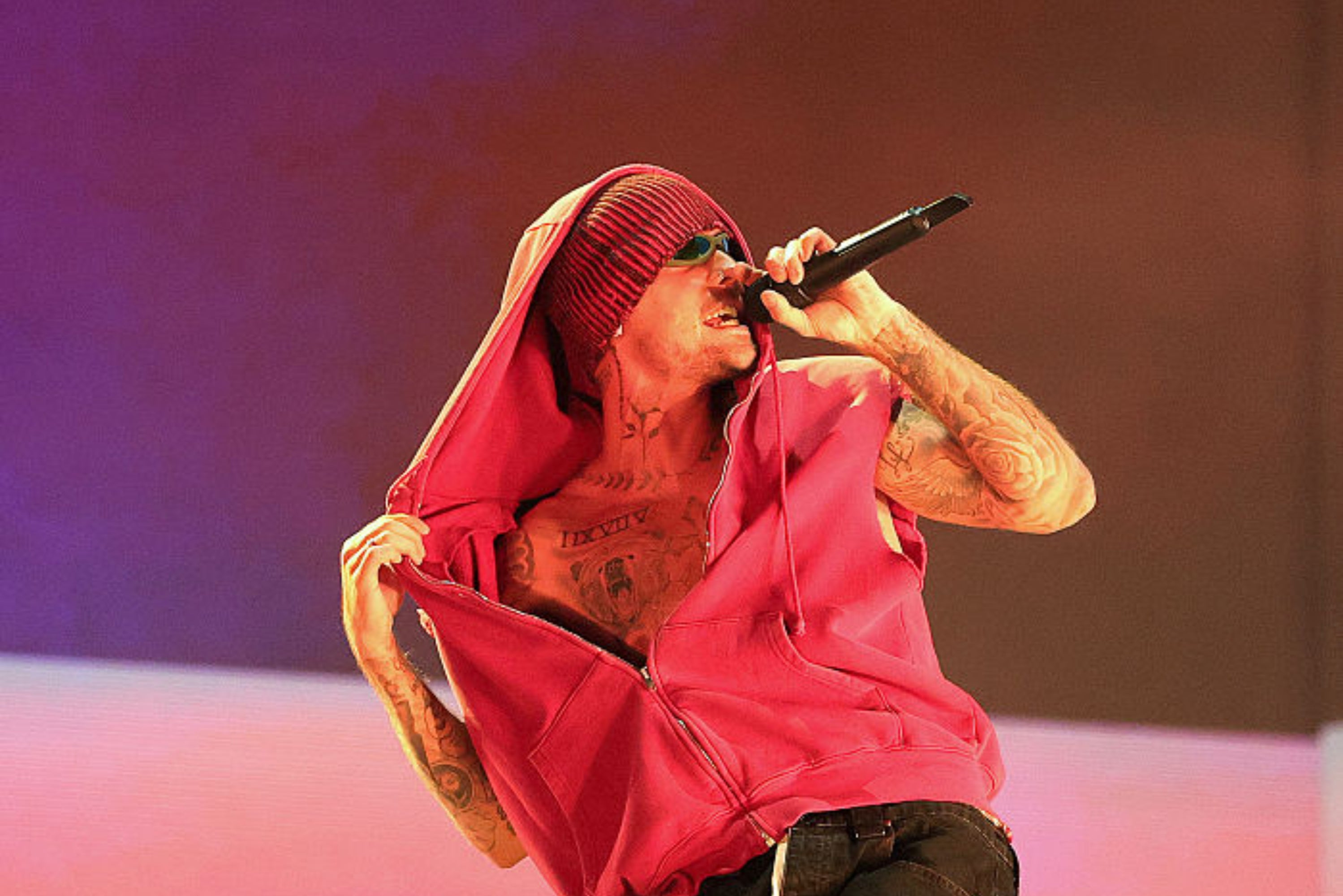 Person performing on stage, wearing a hooded vest and sunglasses. Holding a microphone, showing tattoos while engaging with the audience