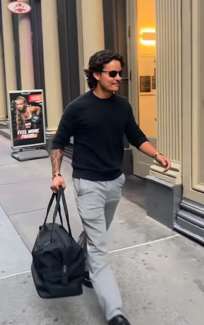 Collin with sunglasses connected  walking connected  a metropolis  street, carrying a achromatic  duffel container  and wearing a achromatic  sweater and airy  pants