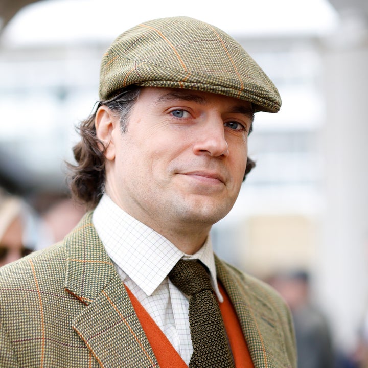 Person in plaid suit and flat cap, looking at the camera. Background shows blurred figures and buildings