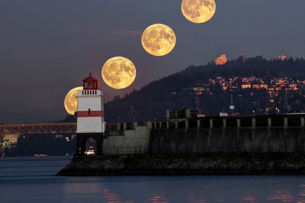 A time-lapse of the full moon rising over a lighthouse and city skyline by the water