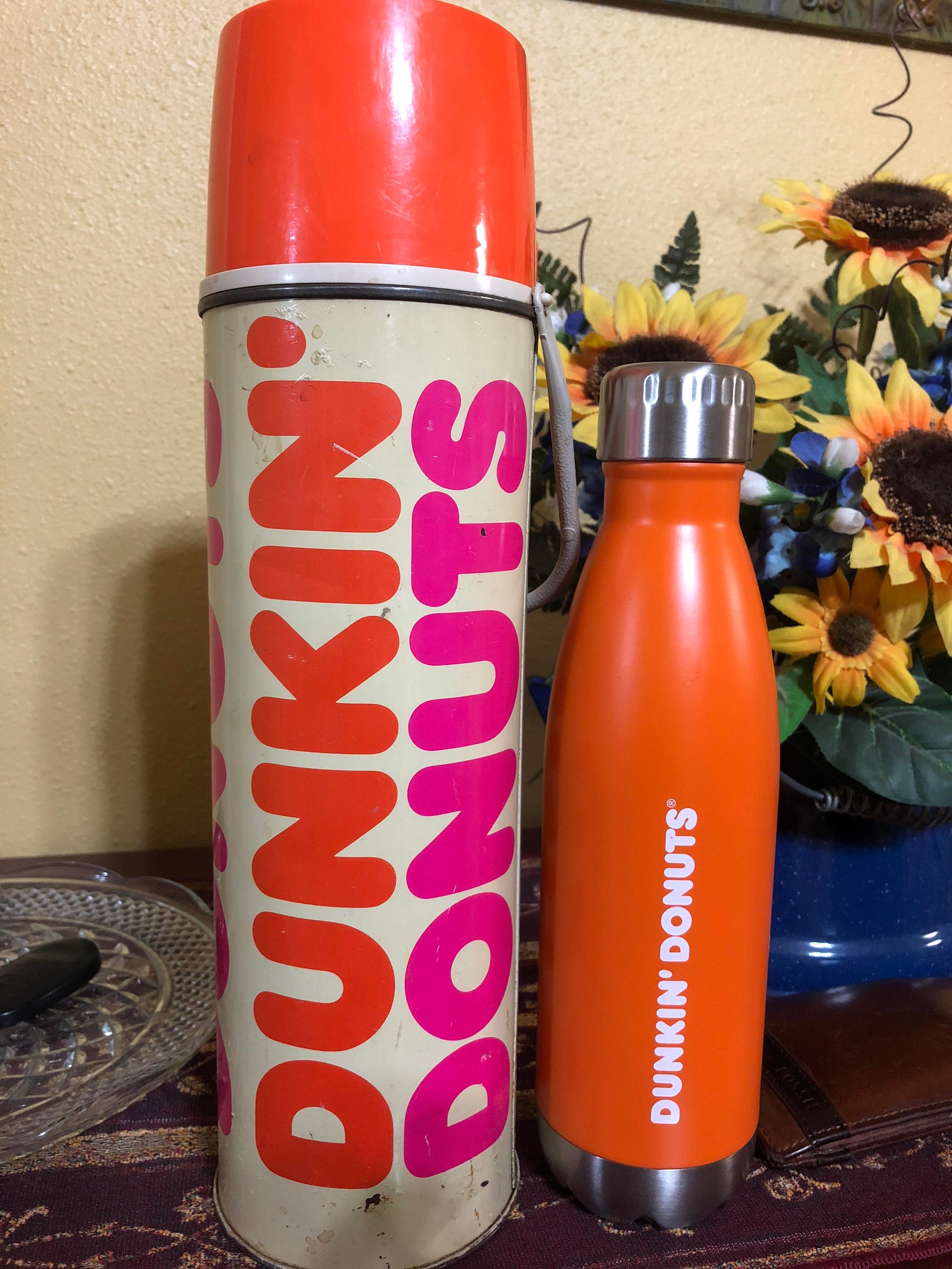 Two Dunkin' Donuts thermoses, vintage and modern, are displayed side by side on a table with sunflowers in the background