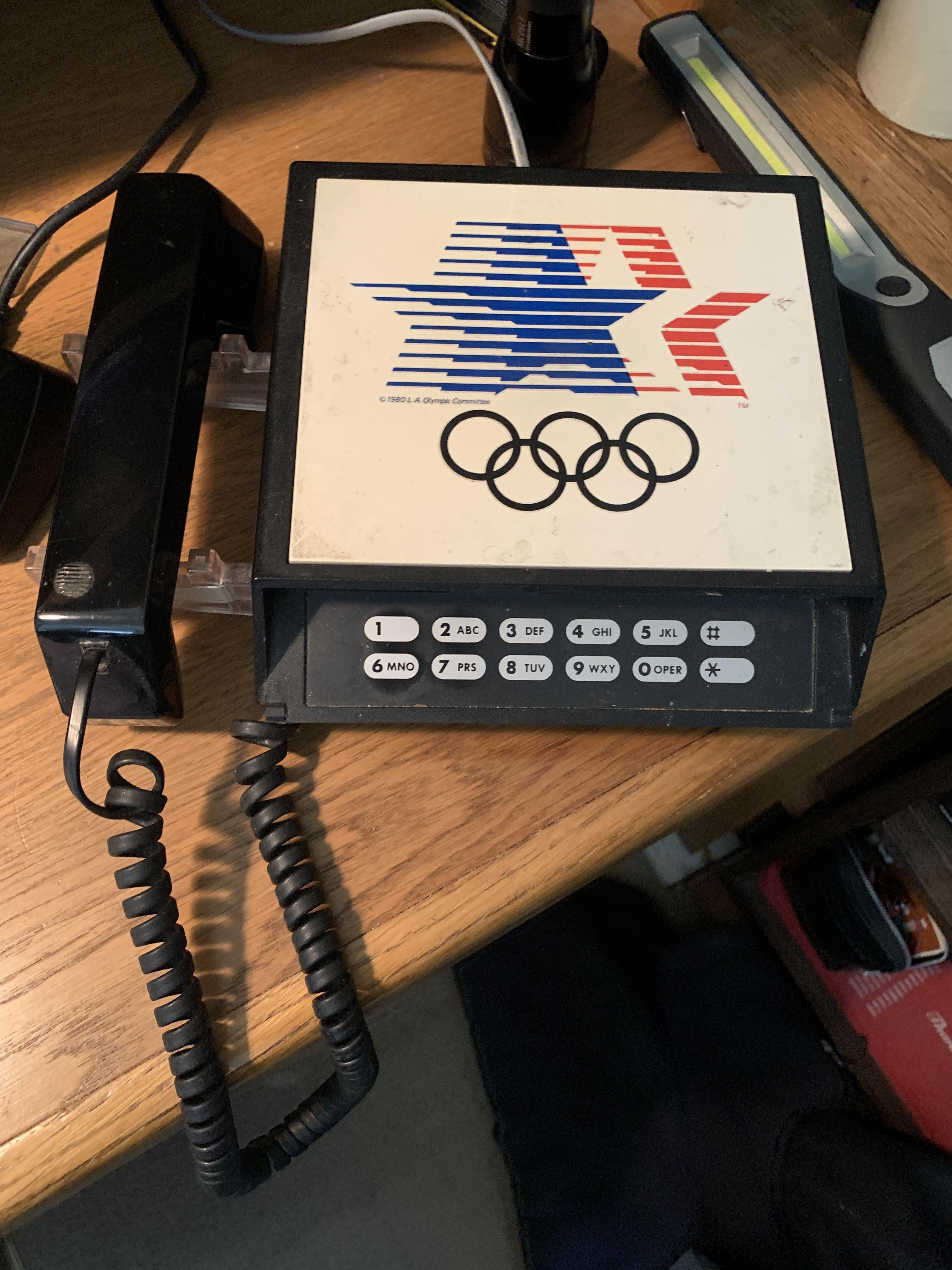 Vintage telephone with Olympic branding and 1984 graphics, featuring a push-button dialing pad and coiled cord on a wooden desk