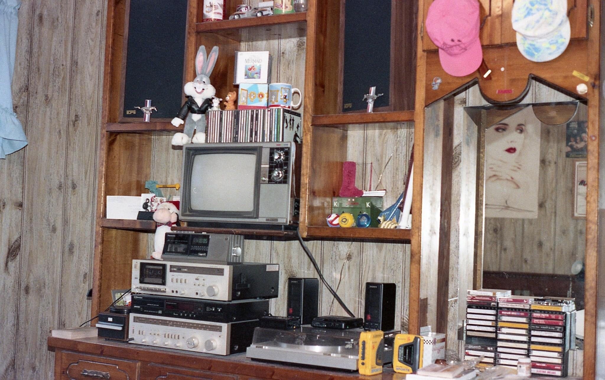 Vintage room with a CRT TV, audio equipment, shelves with cassettes and CDs, and decorative items. A mirror reflects part of a poster