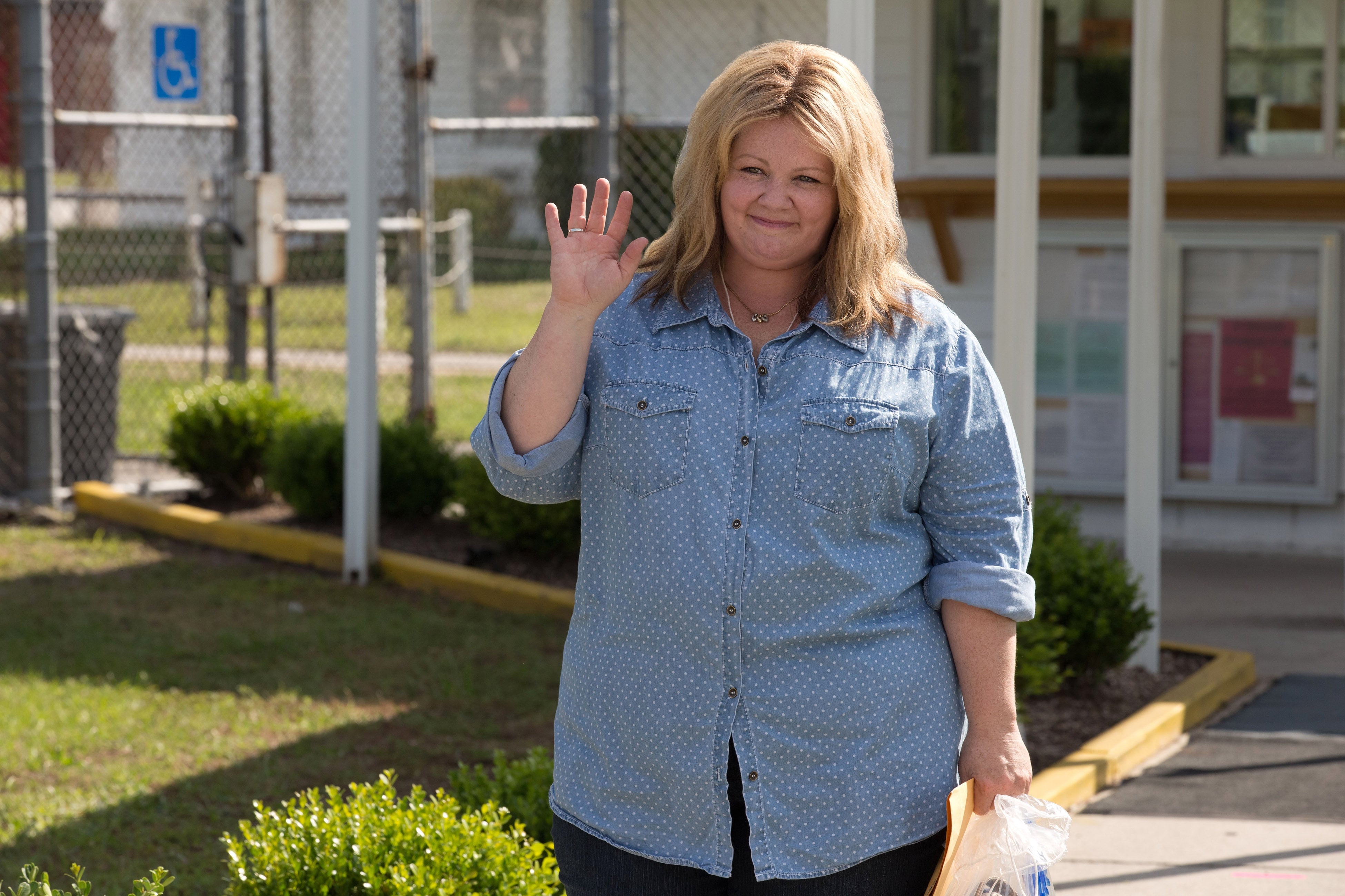 Melissa McCarthy with a denim shirt waving and holding a paper bag outside a building, smiling at the camera