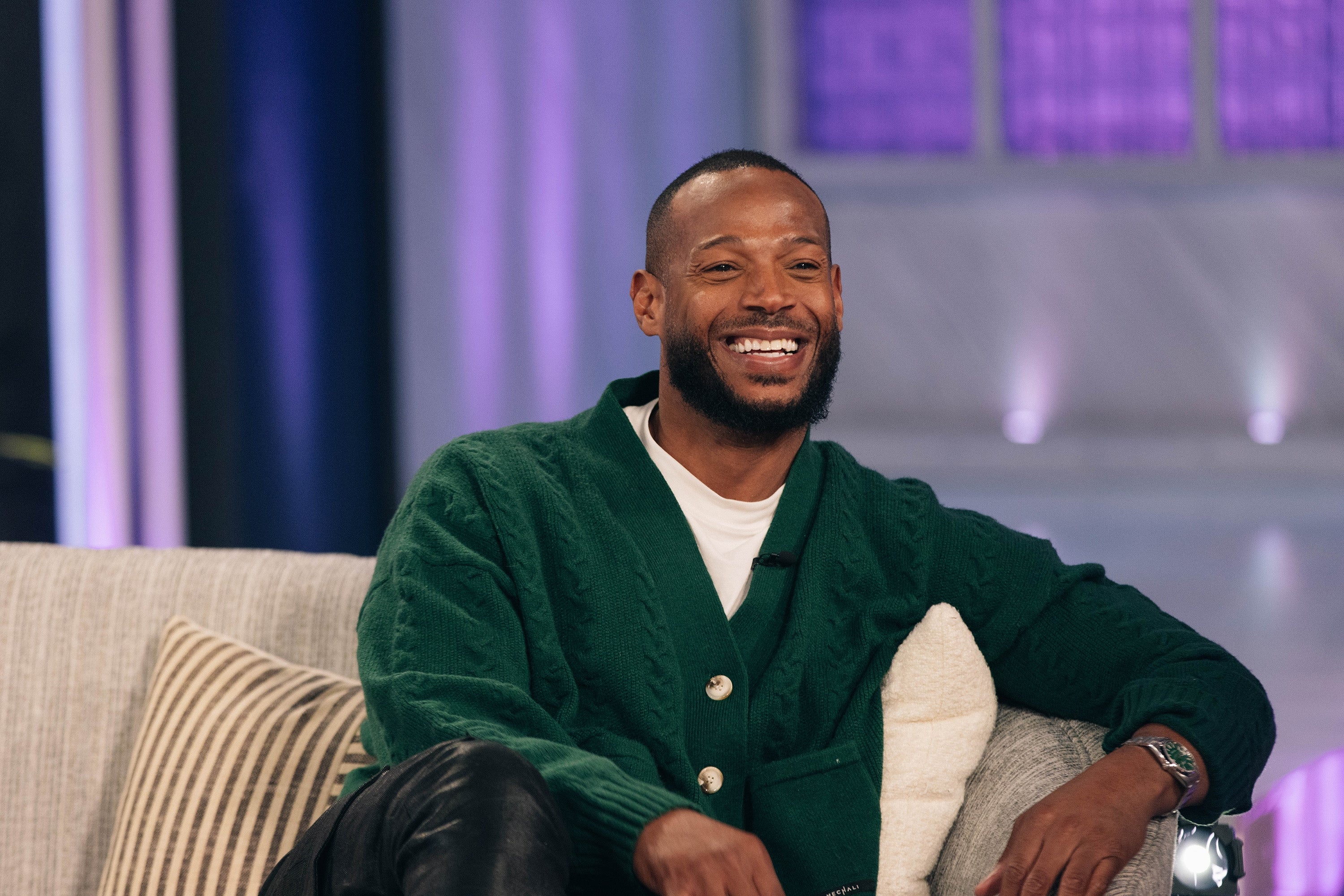 Marlon Wayans in a cardigan sits smiling on a couch during a talk show appearance
