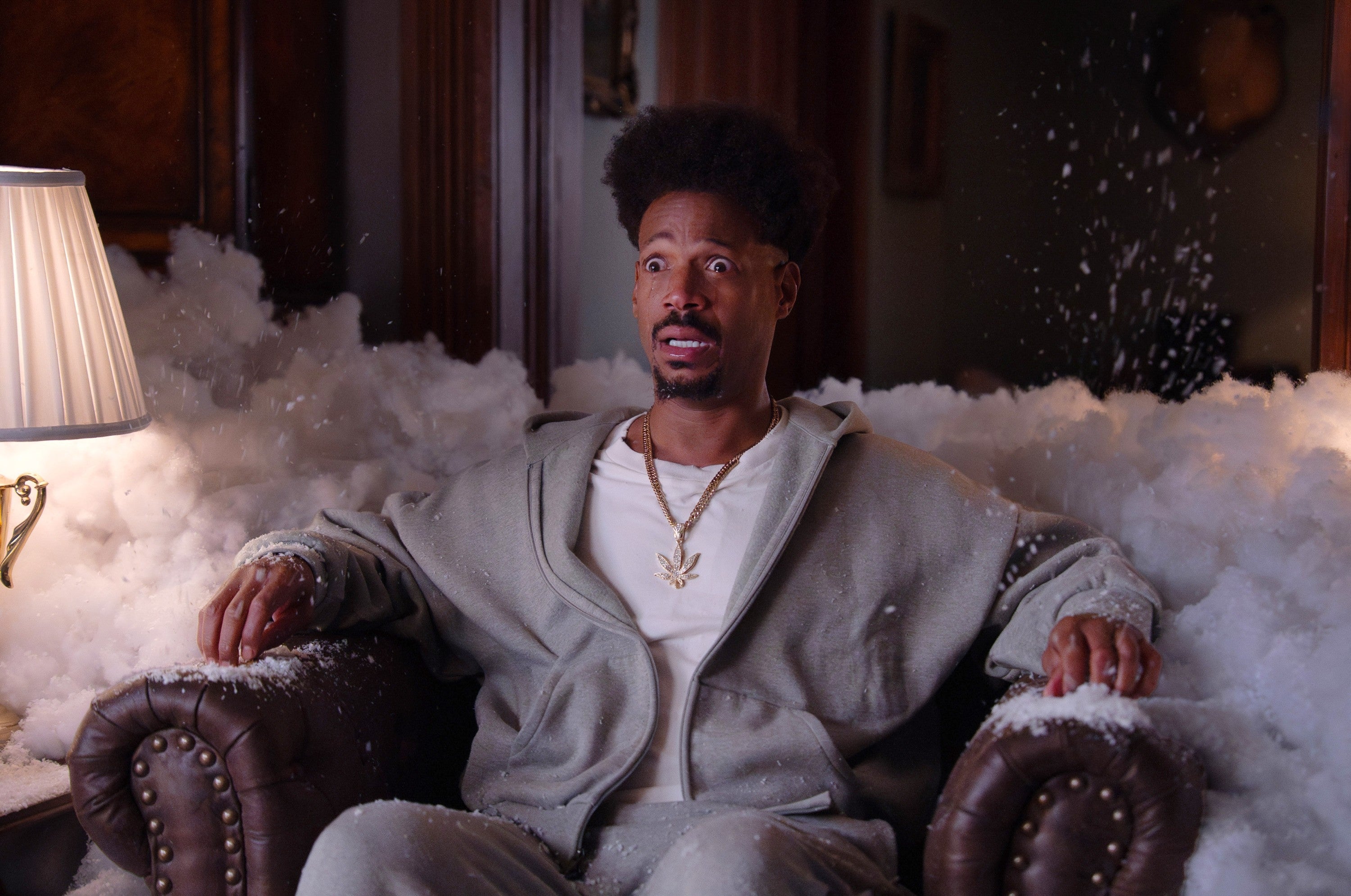Marlon Wayans sits surprised on a couch surrounded by soap bubbles, wearing a casual sweater and pants, with a lamp nearby