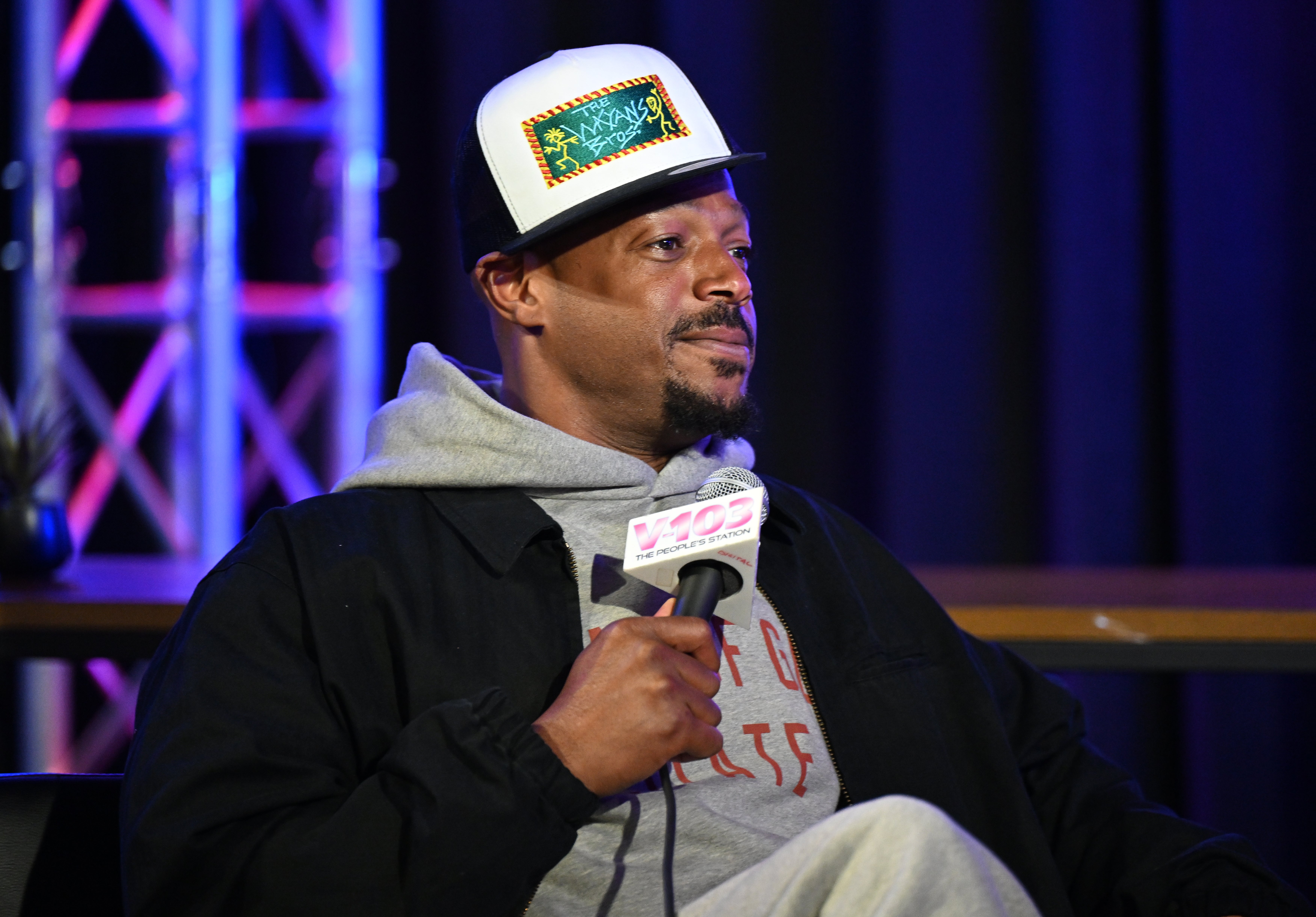 Marlon Wayans at a media event holding a microphone, wearing a hoodie and a cap