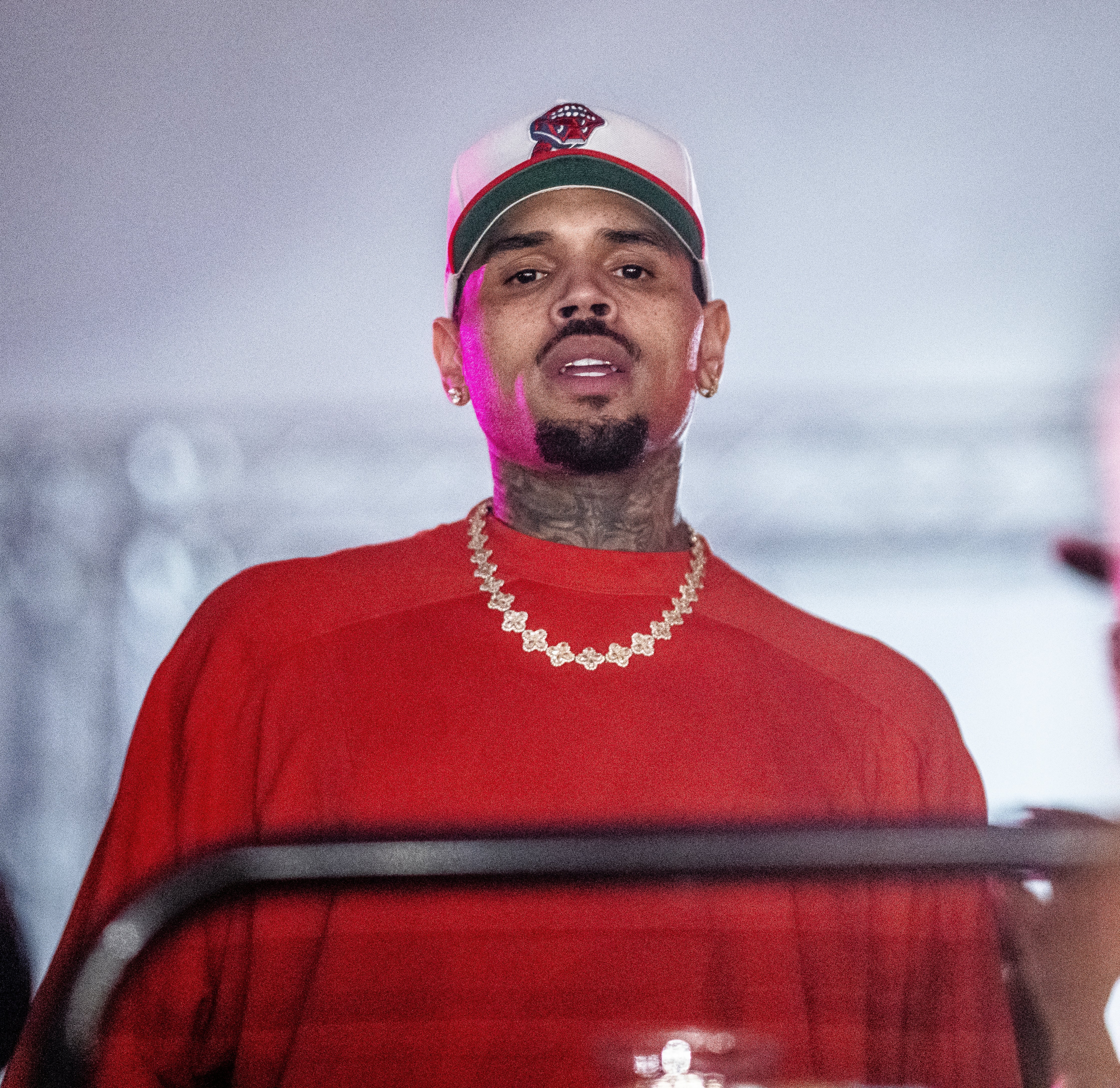 Chris Brown wearing a baseball cap and chain necklace, looking directly ahead, in a casual setting