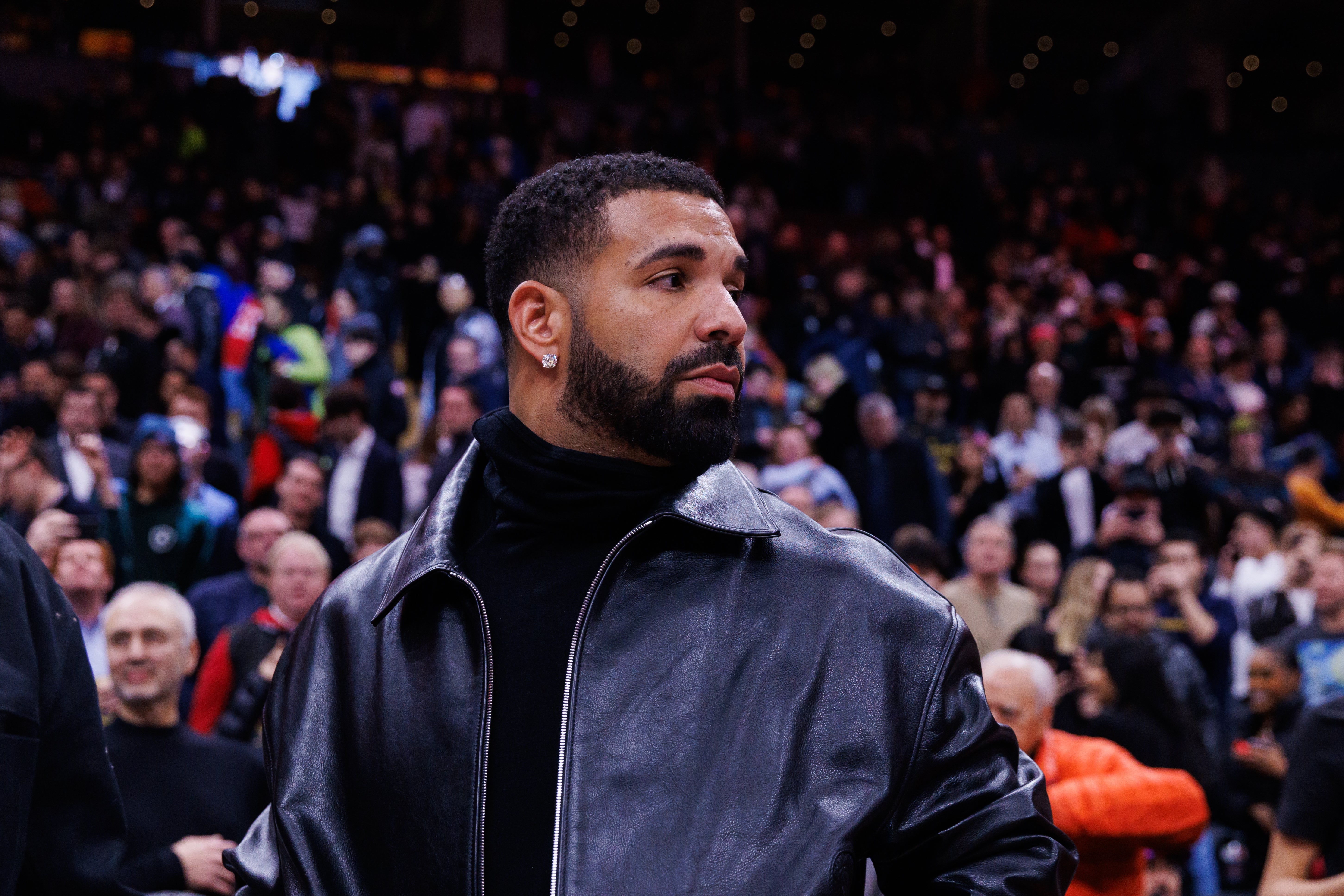 Drake in a leather jacket and turtleneck stands in a crowded arena, looking to the side