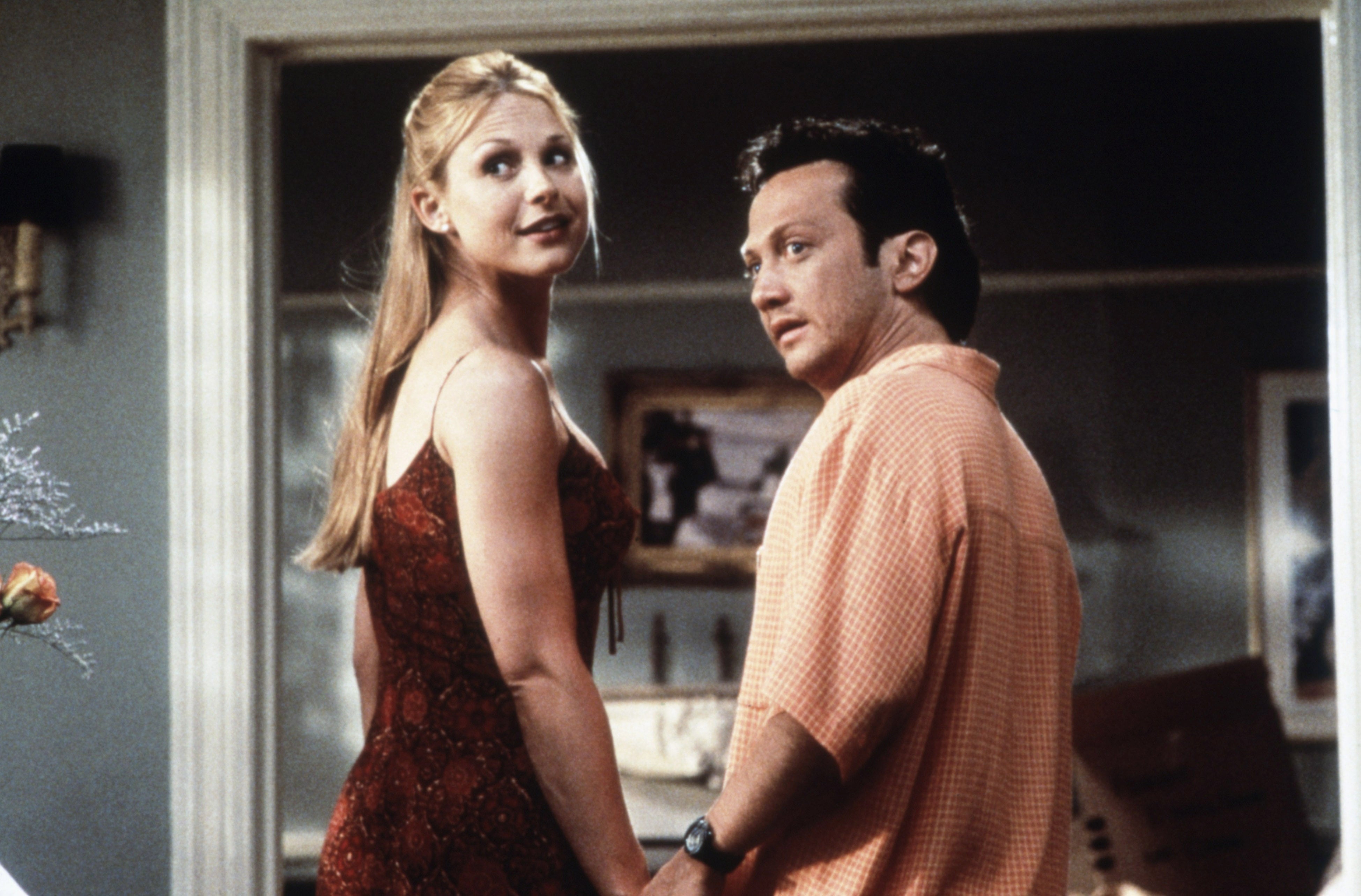 Arija Bareikis and Rob Schneider stand indoors, holding hands in a scene from Deuce Bigalow Male Gigolo