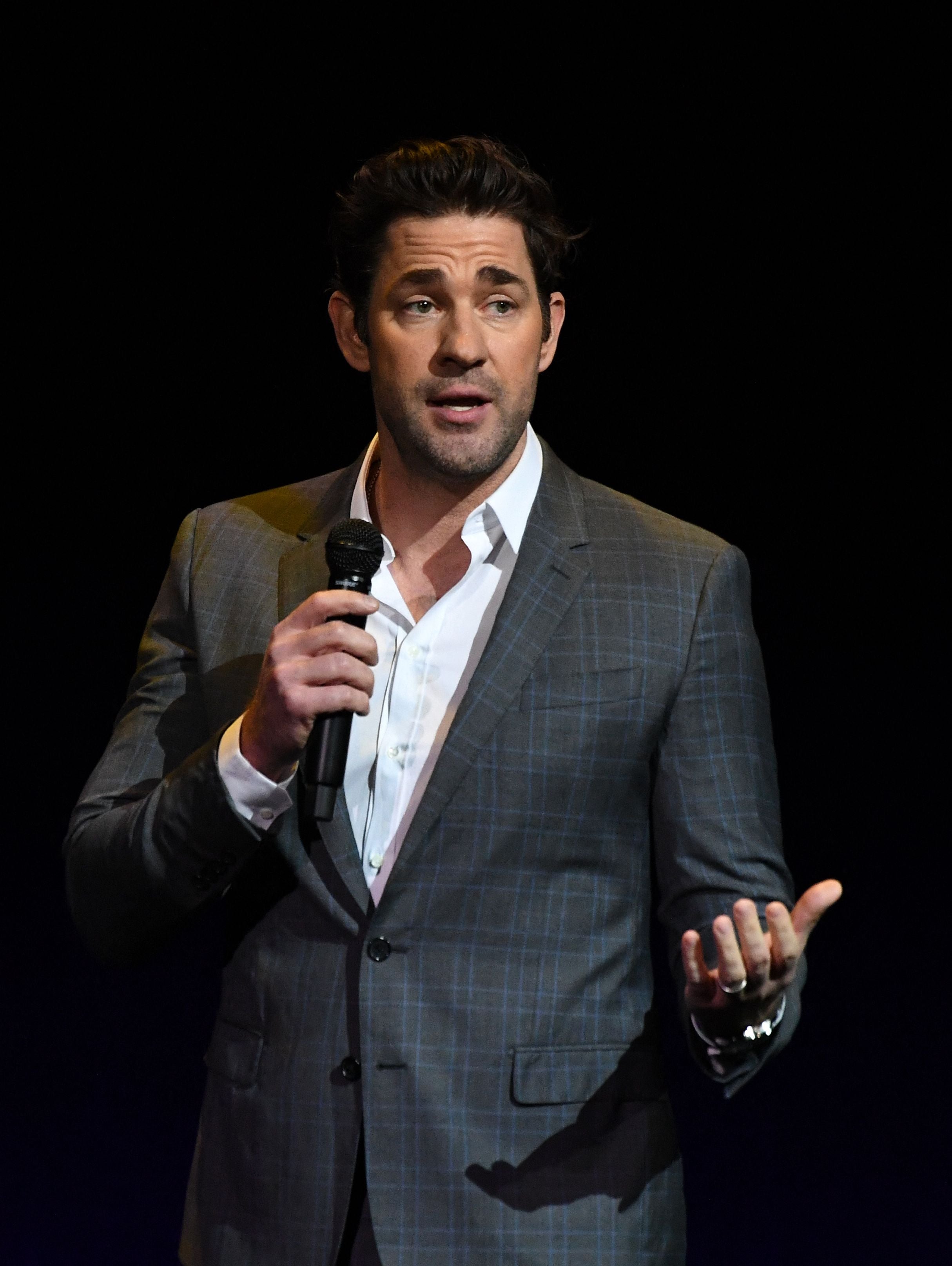 John Krasinski in a checkered suit and shirt speaks into a microphone on stage, gesturing with one hand