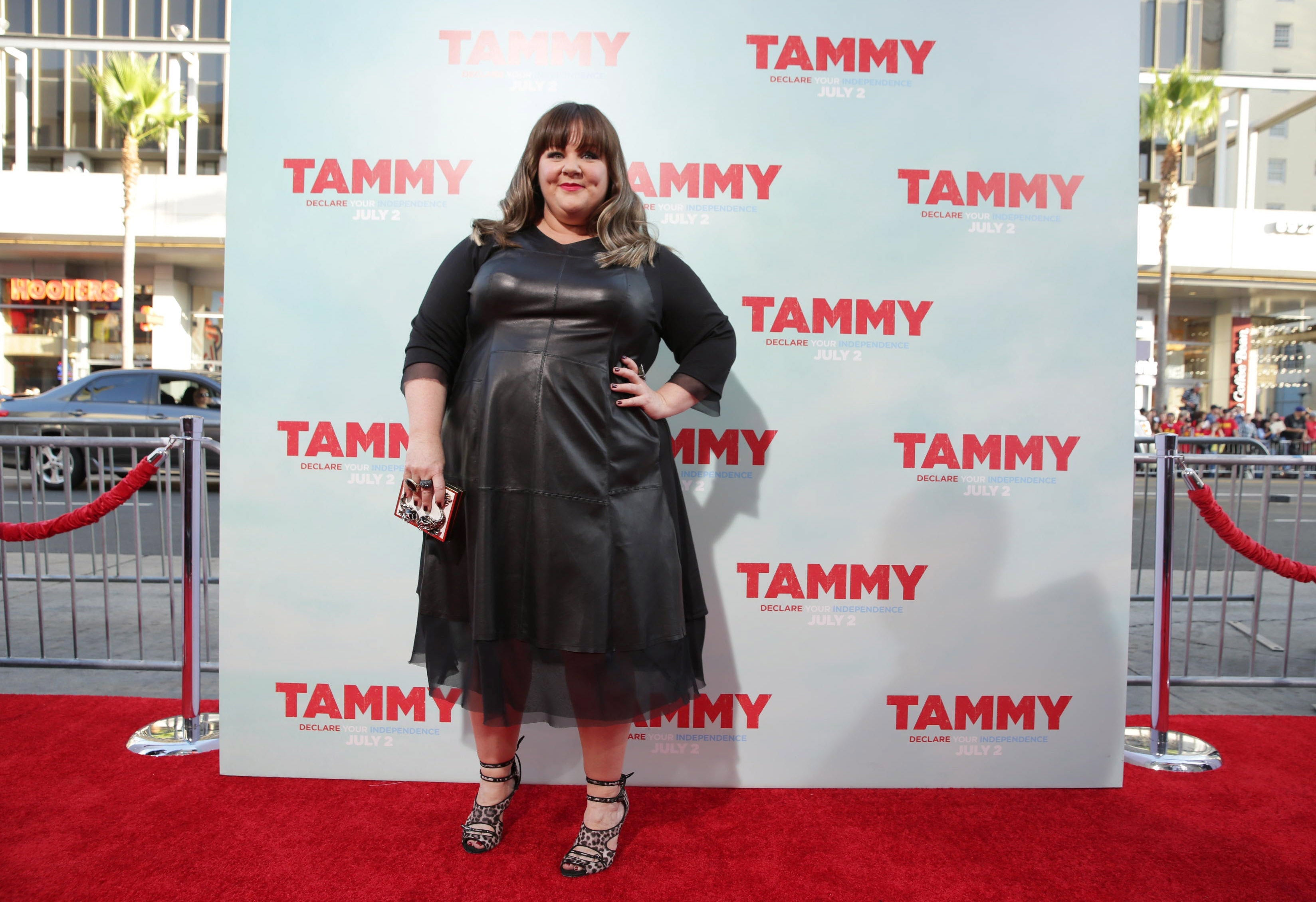 Melissa McCarthy posing on a red carpet, wearing a sheer dress and holding a clutch, at an event with a "Tammy" backdrop