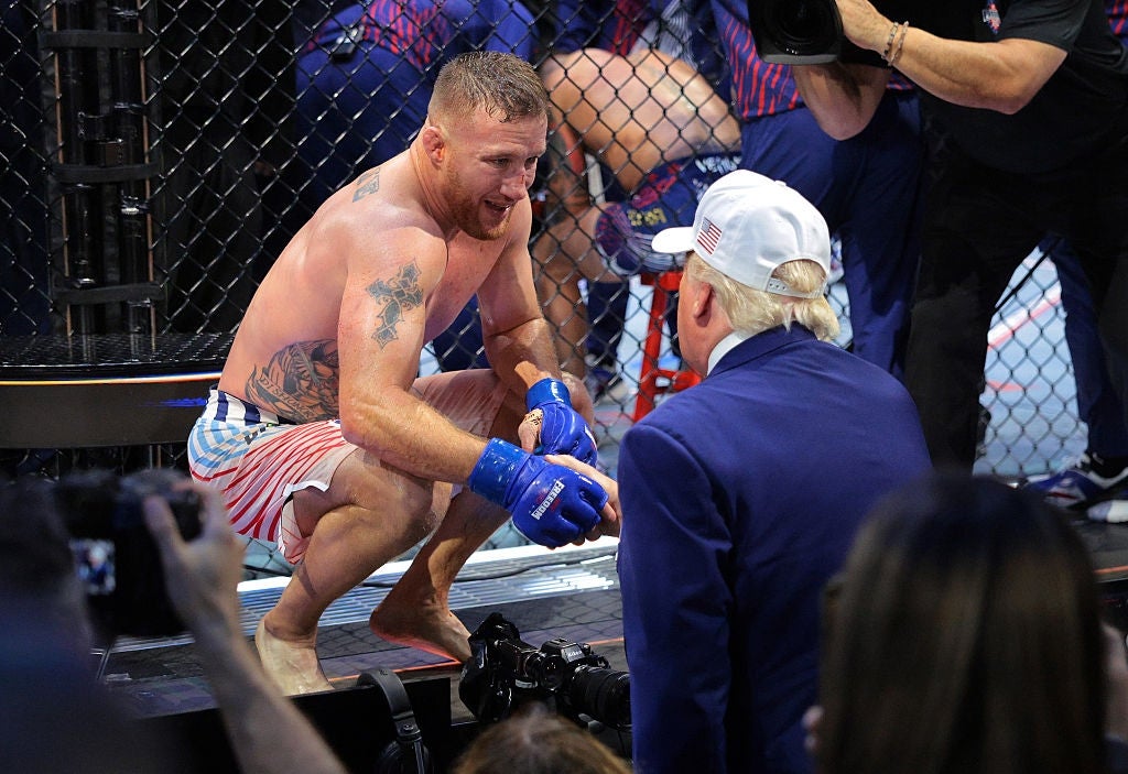 MMA fighter sitter på huk inne i et bur, snakker til en mann i dress og caps utenfor buret, omringet av mediepersonell