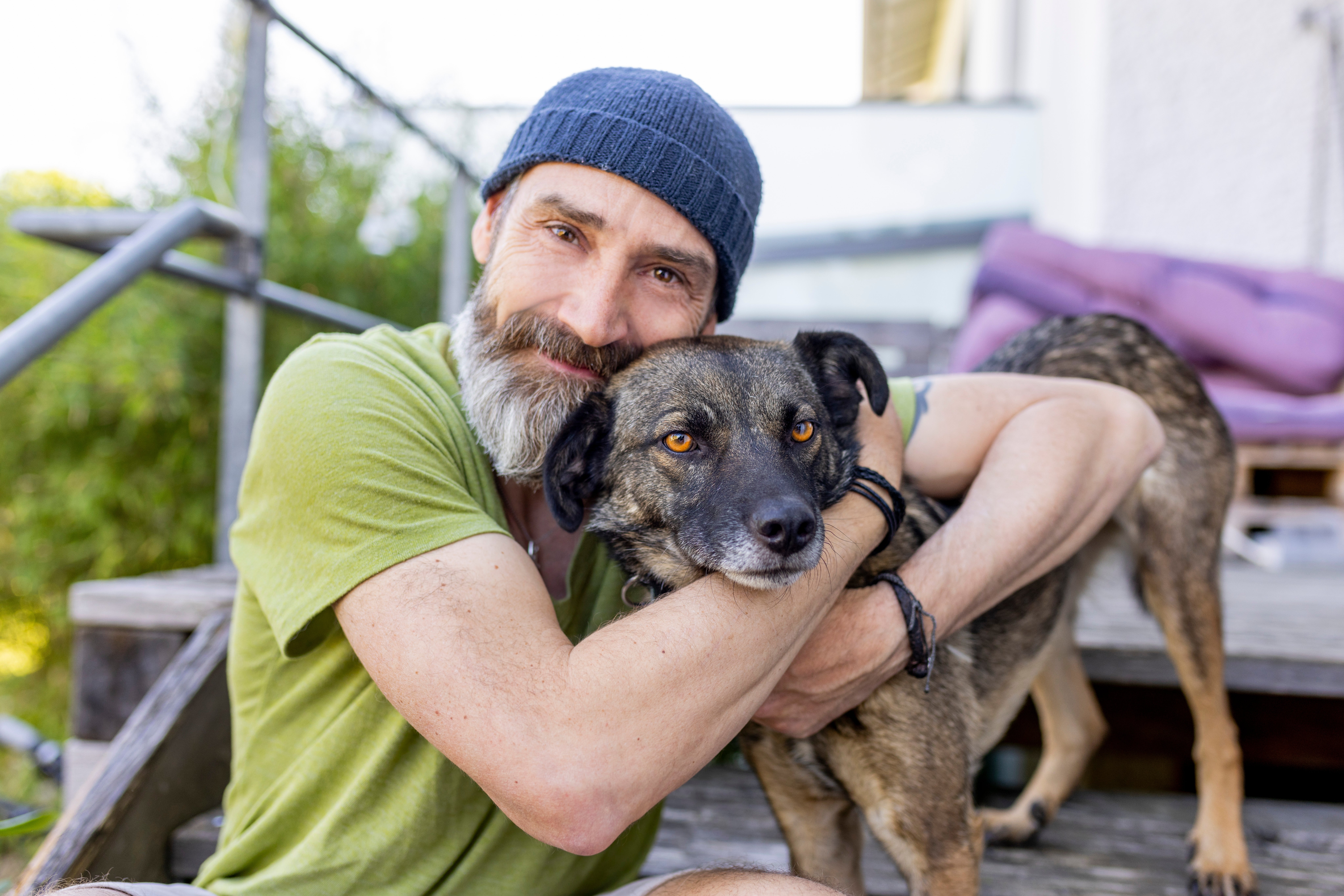 Man with a beard wearing a beanie and greenish  t-shirt, sitting connected  steps, lovingly hugging a brownish  and achromatic  dog