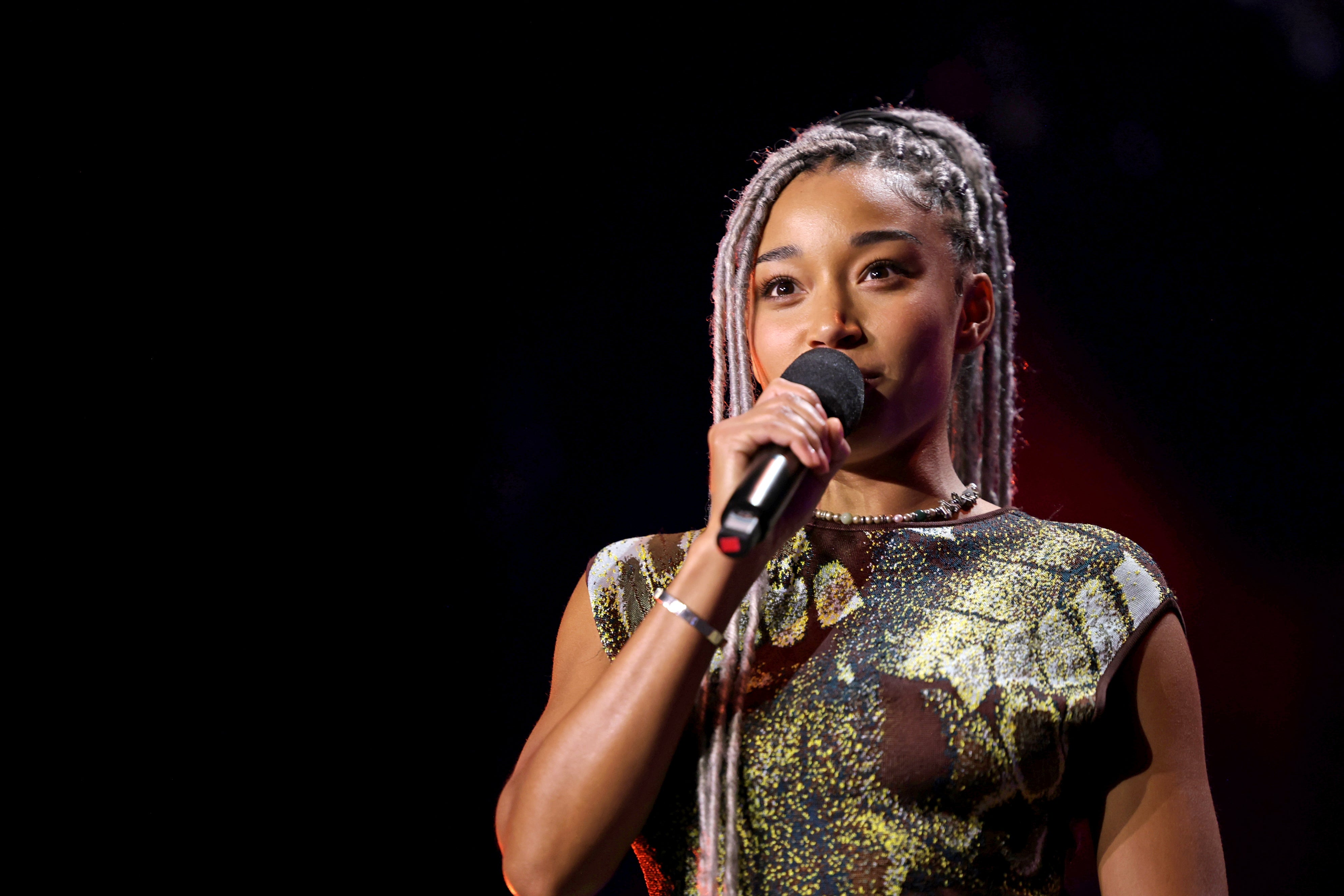Amandla Stenberg with braided hair holds a microphone, wearing a stylish patterned dress, against a plain background