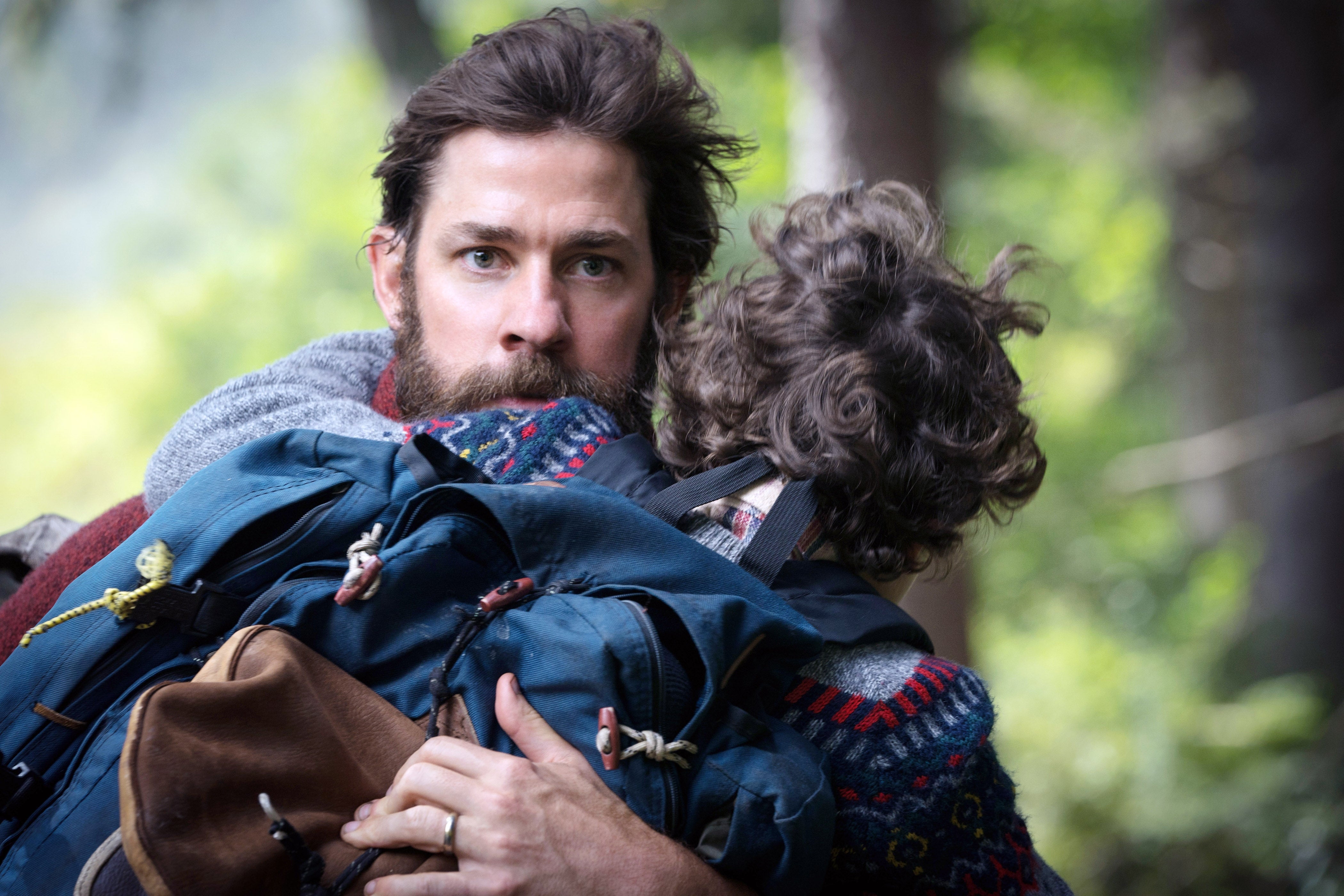 John Krasinski with a beard carries Noah Jupe through a forest, looking alert in a scene from A Quiet Place
