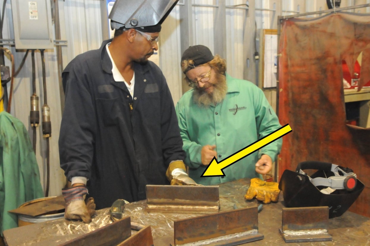 Two people in a workshop discussing metalworking, with a yellow arrow pointing to a piece of metal they are examining