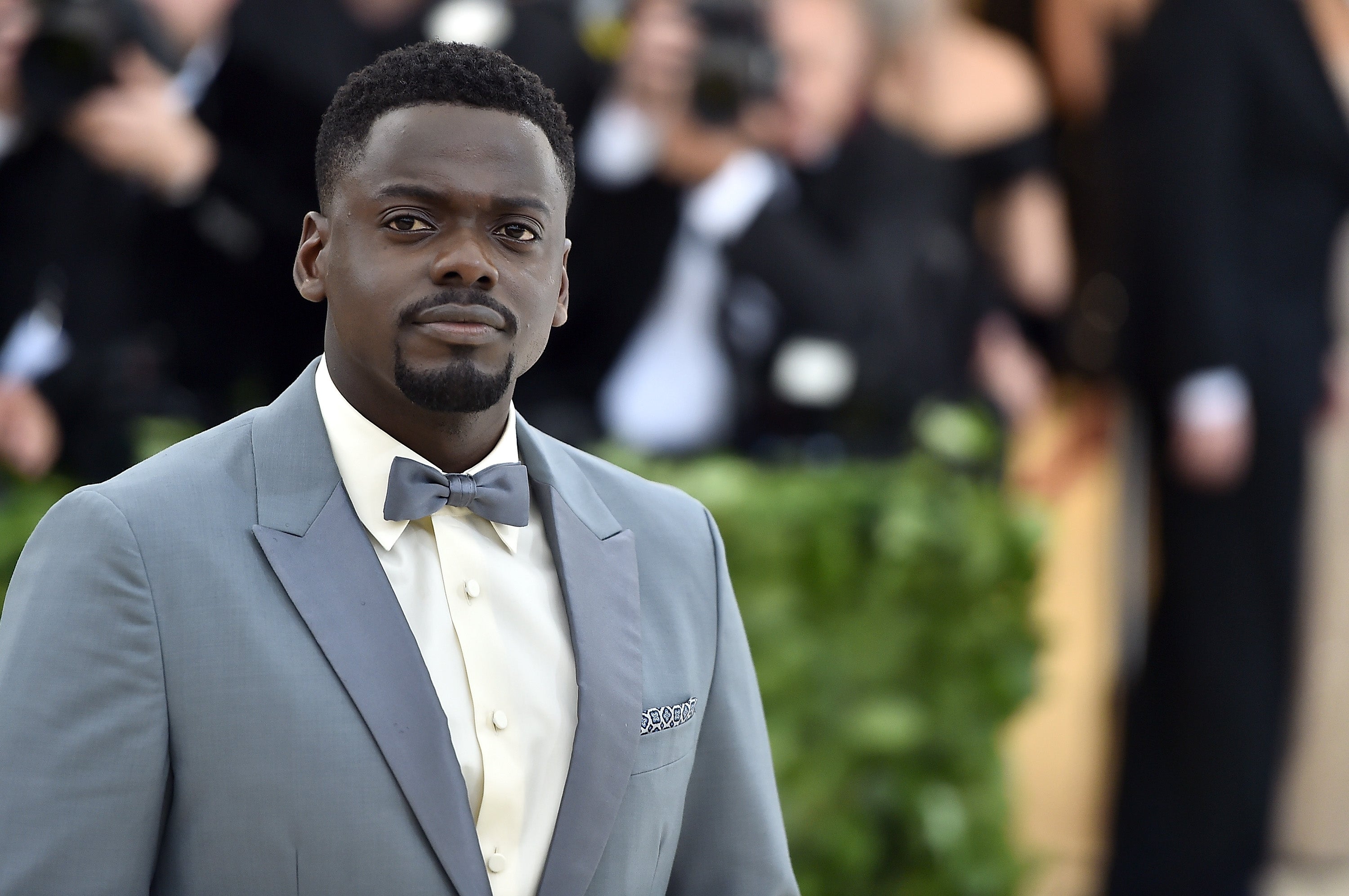 Daniel Kaluuya in a formal suit and bow tie stands on a red carpet, surrounded by blurred attendees in the background