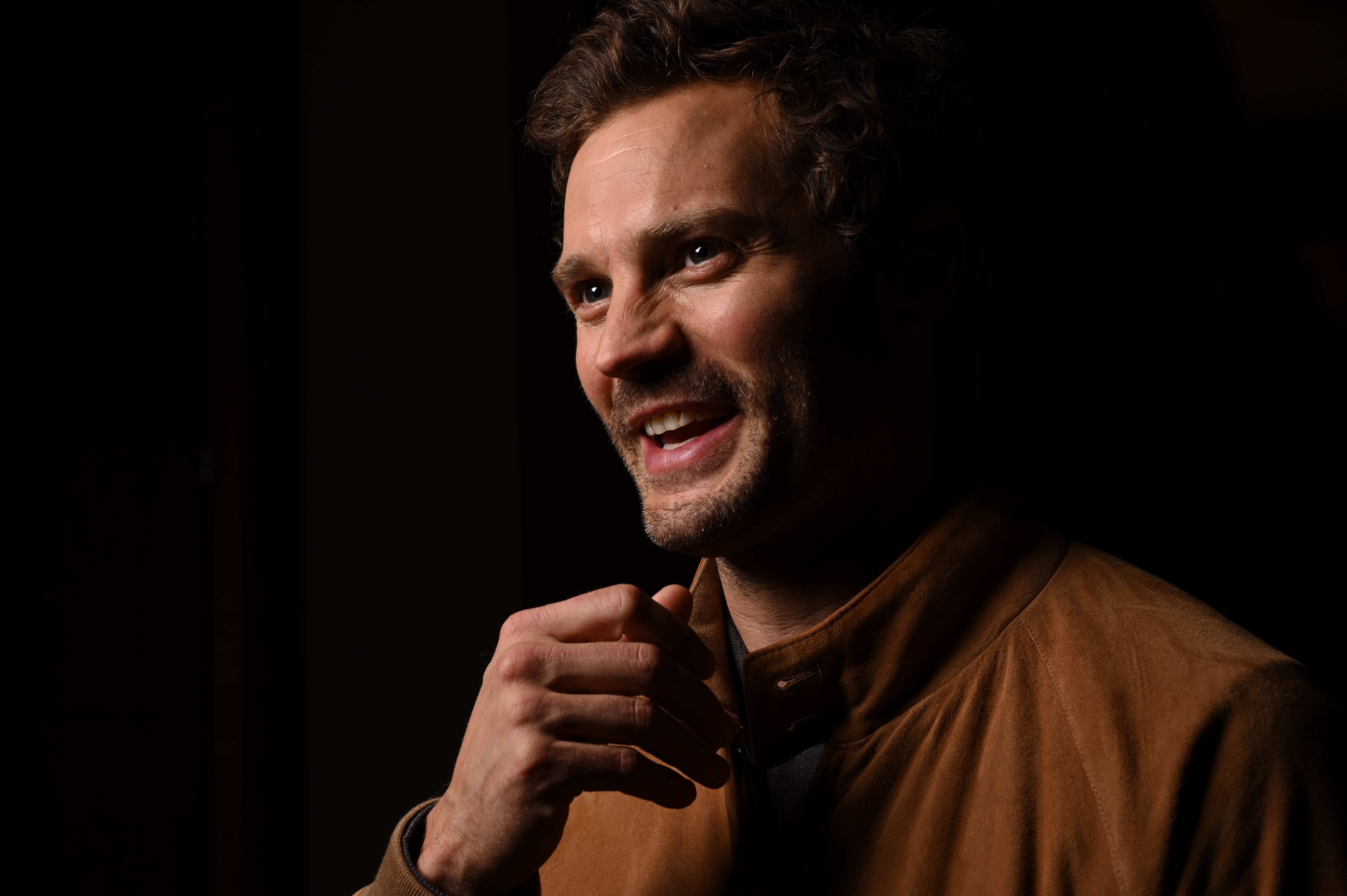 Jamie Dornan smiling, wearing a casual jacket, looking off-camera in a dark setting
