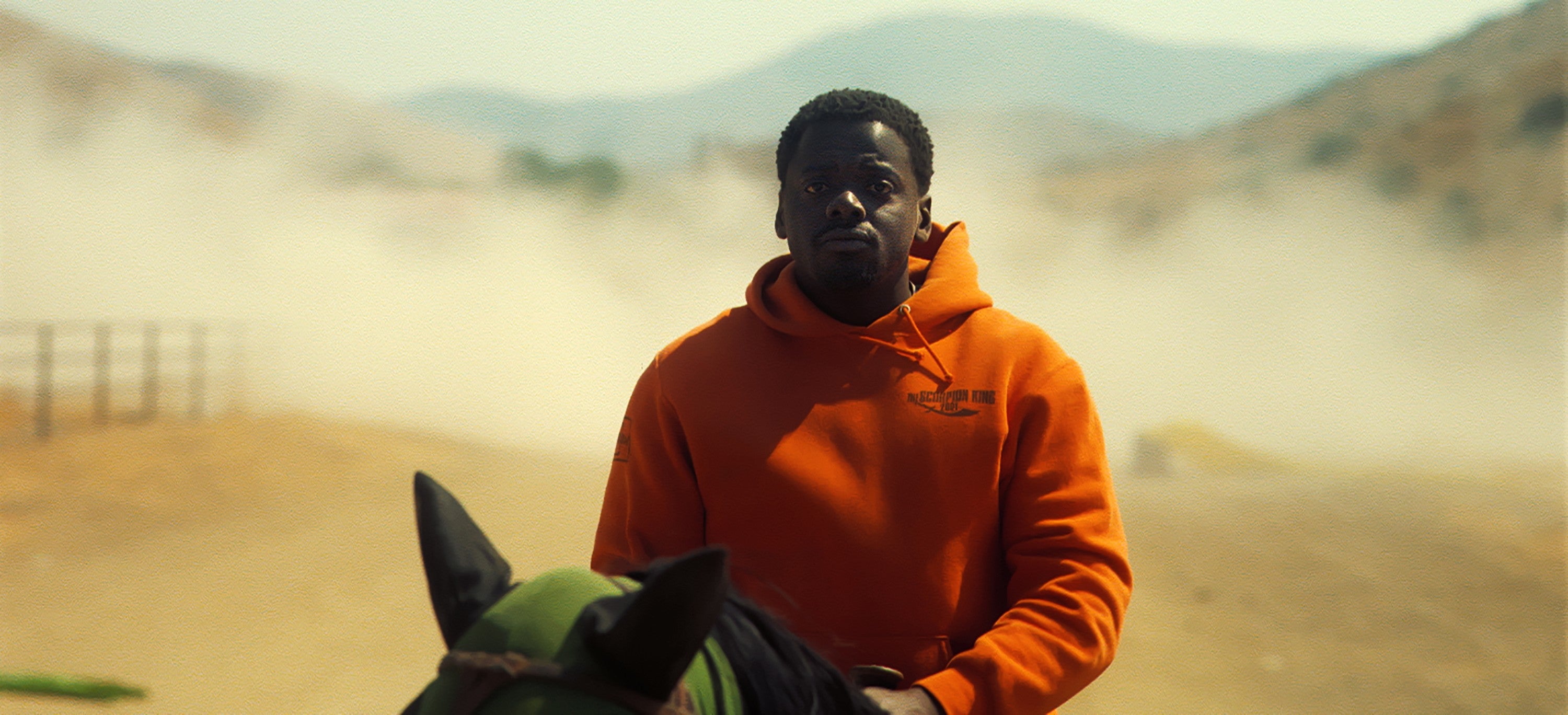 Daniel Kaluuya riding a horse on a dusty road, wearing a casual hoodie, against a mountainous backdrop in a scene from Nope