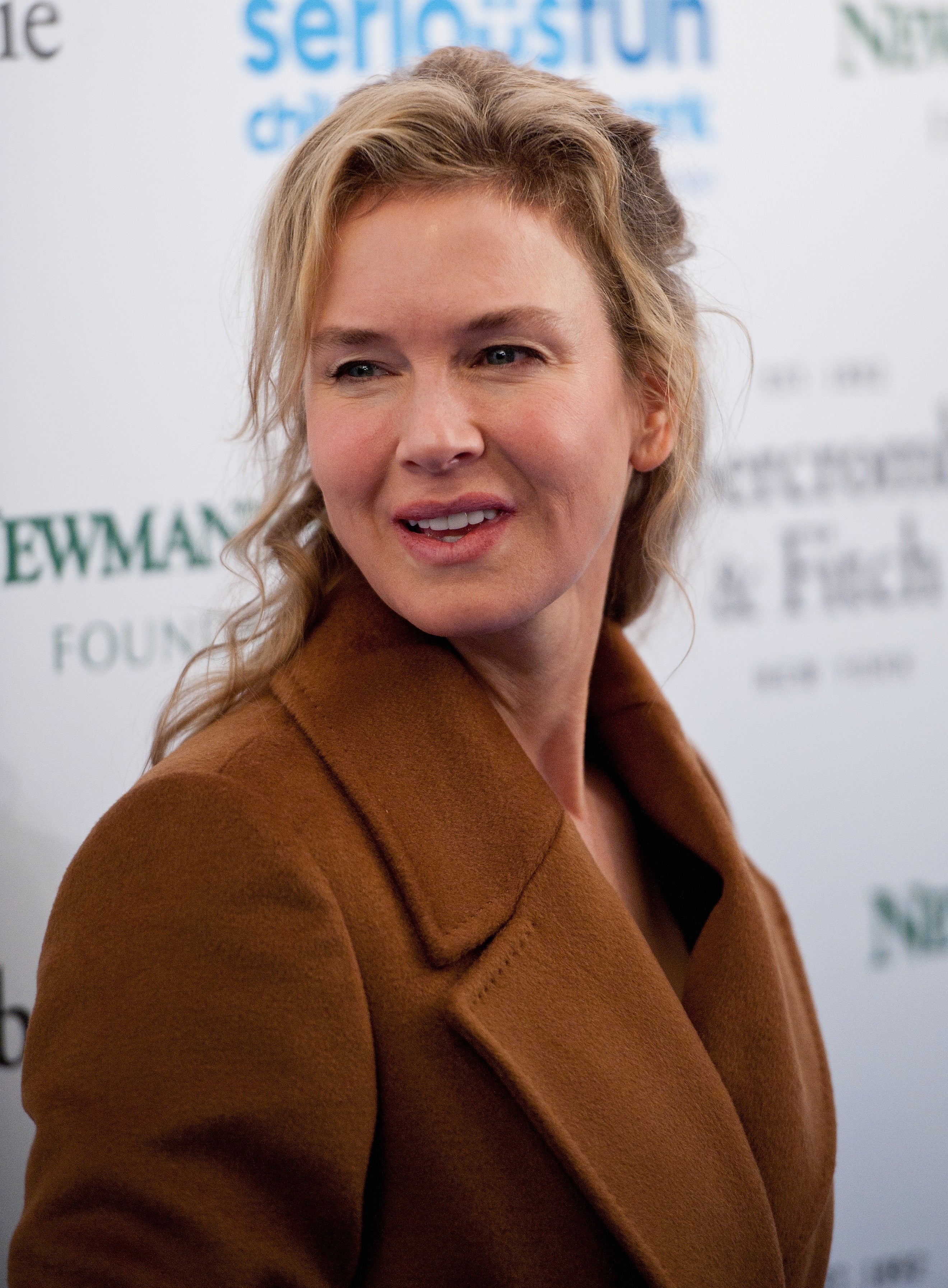 Renee Zellwegger wearing a coat, posing at a celebrity event backdrop with logos and text