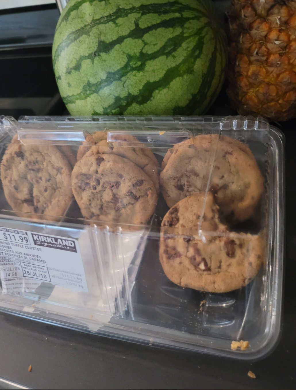 Pack of chocolate chip cookies with one half-eaten, placed next to a watermelon and pineapple on a counter