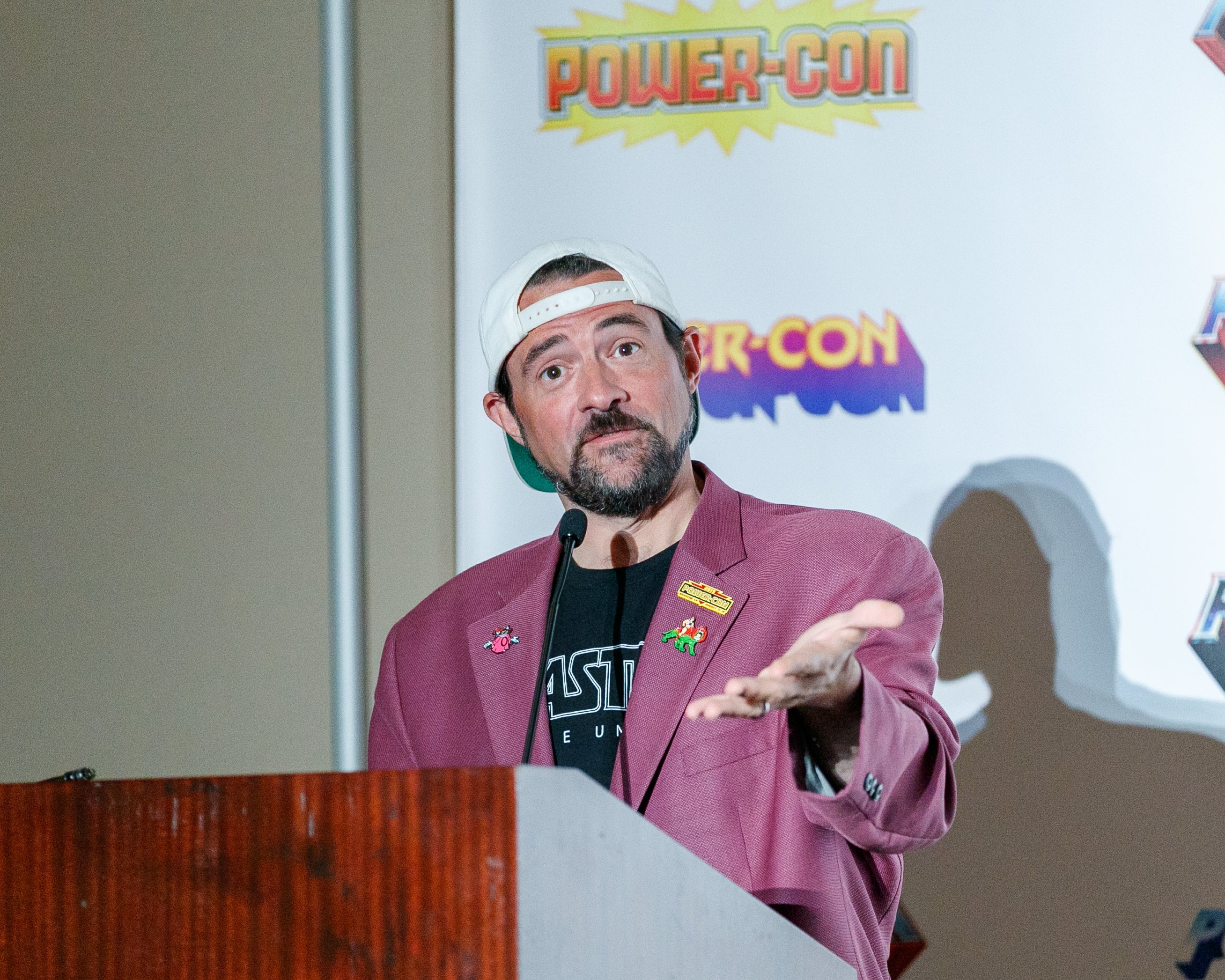 Kevin Smith in a casual blazer and cap speaks at Power-Con event podium
