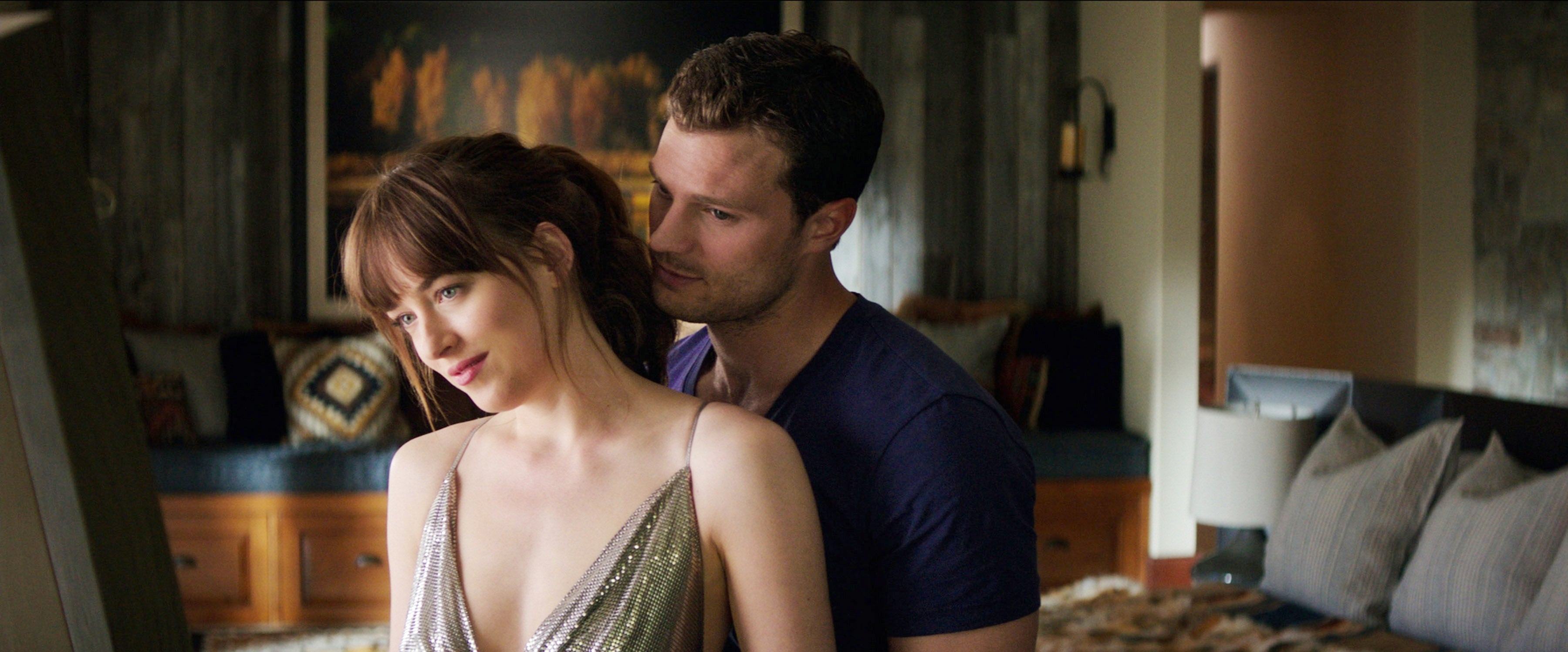 Dakota Johnson and Jamie Dornan stand closely in a cozy room in a scene from Fifty Shades of Grey