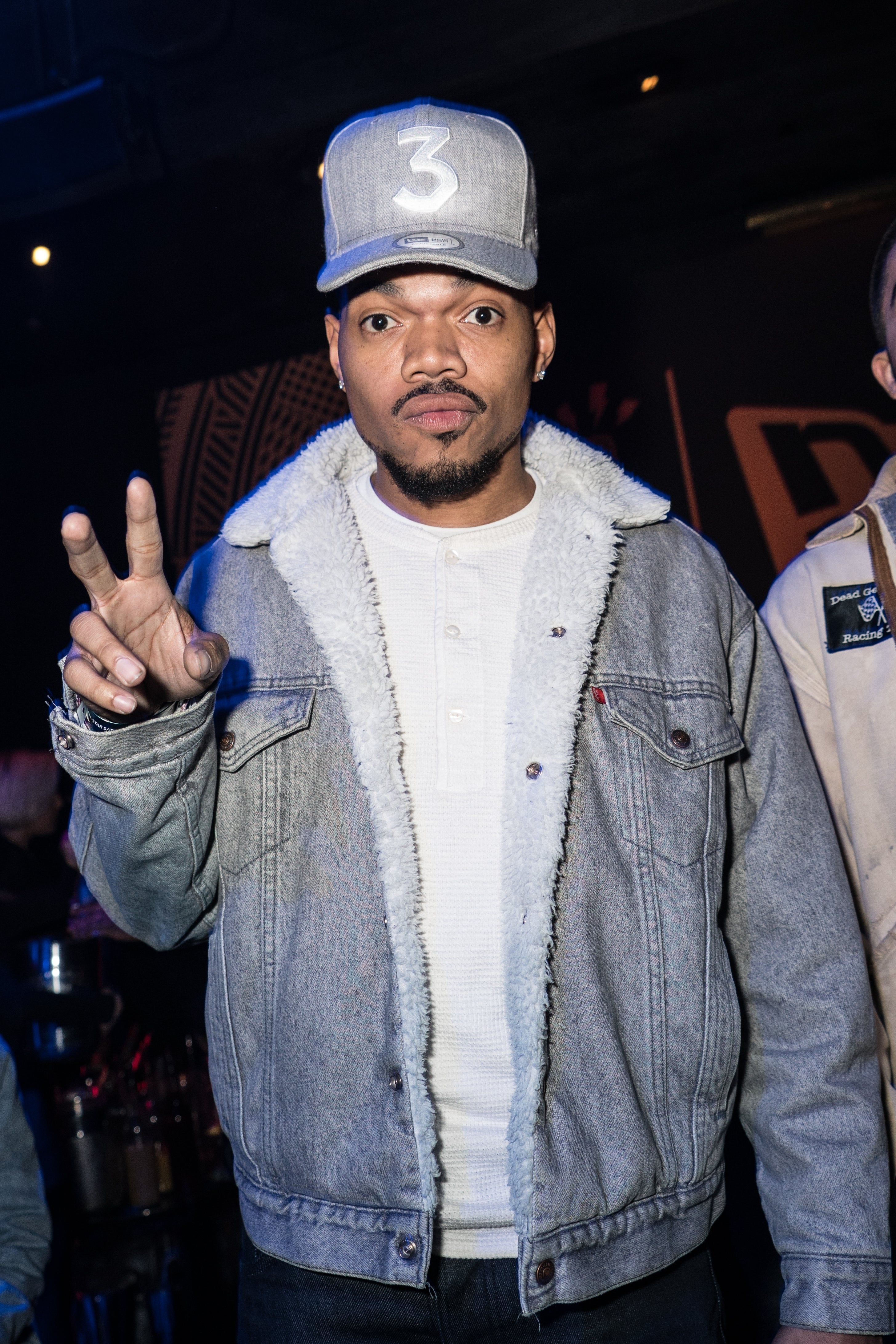 Chance the Rapper in casual denim jacket and cap flashes a peace sign at an event
