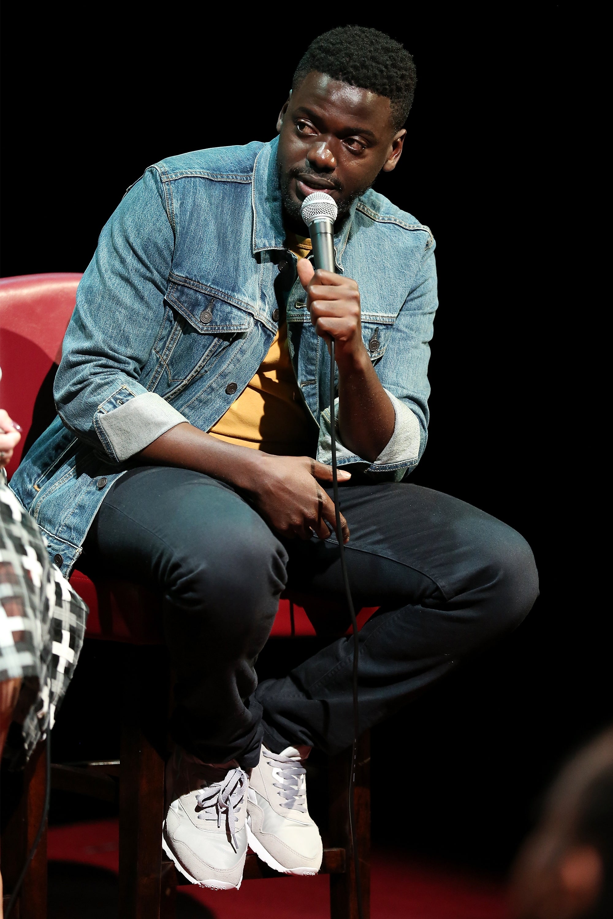 Daniel Kaluuya seated on stage, holding a microphone, wearing a denim jacket and sneakers