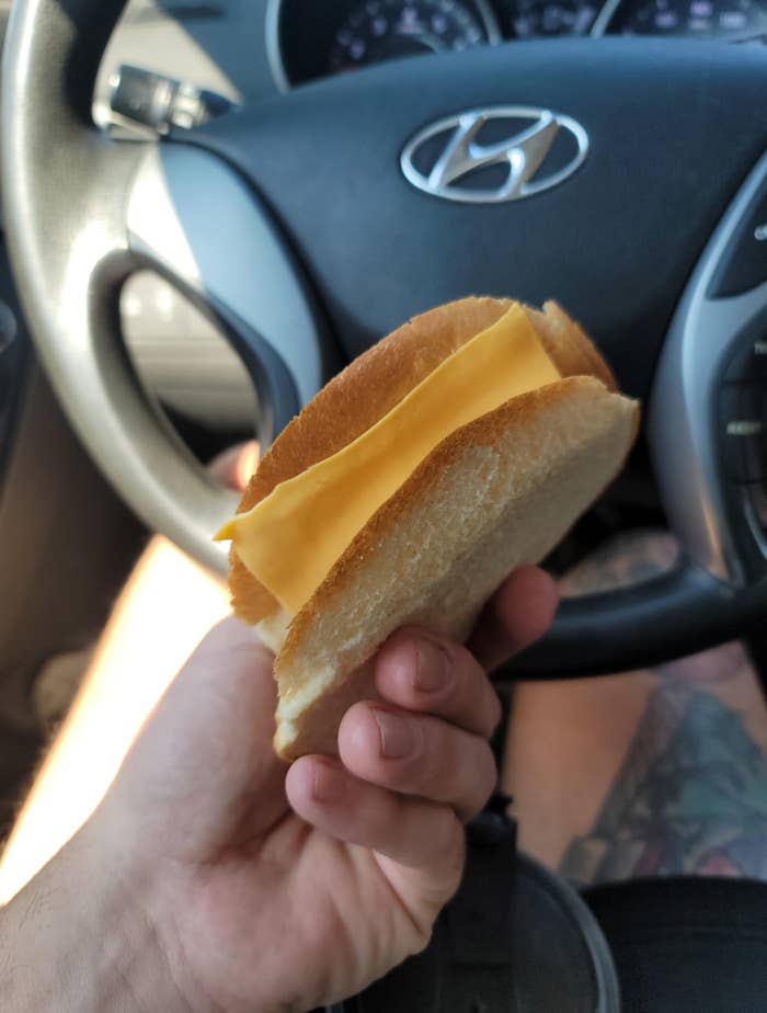 Hand holding a bland cheese sandwich inside a car, with the steering wheel visible in the background