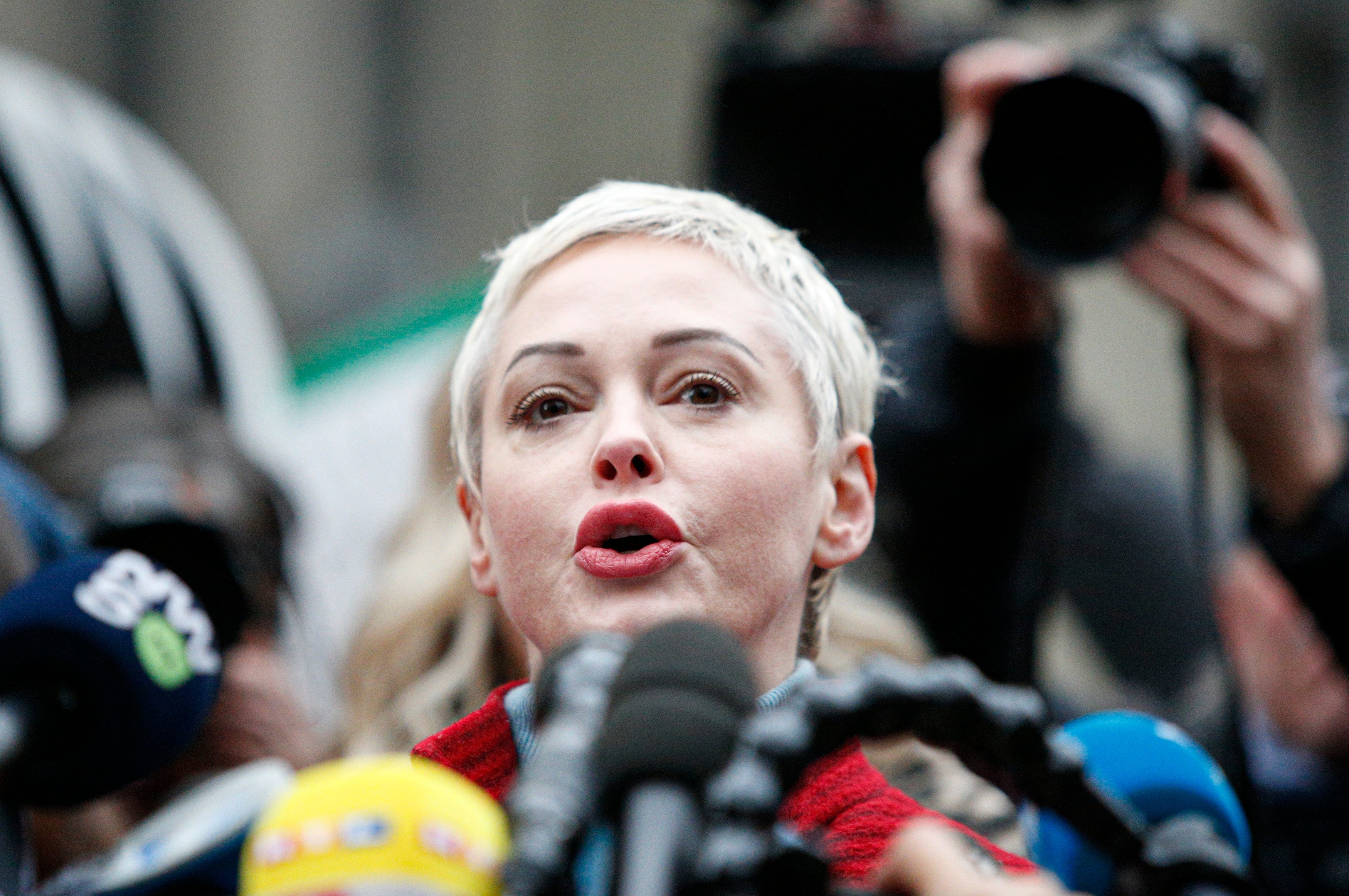 Rose McGowan with short hair speaking into microphones at a press event, surrounded by cameras and reporters