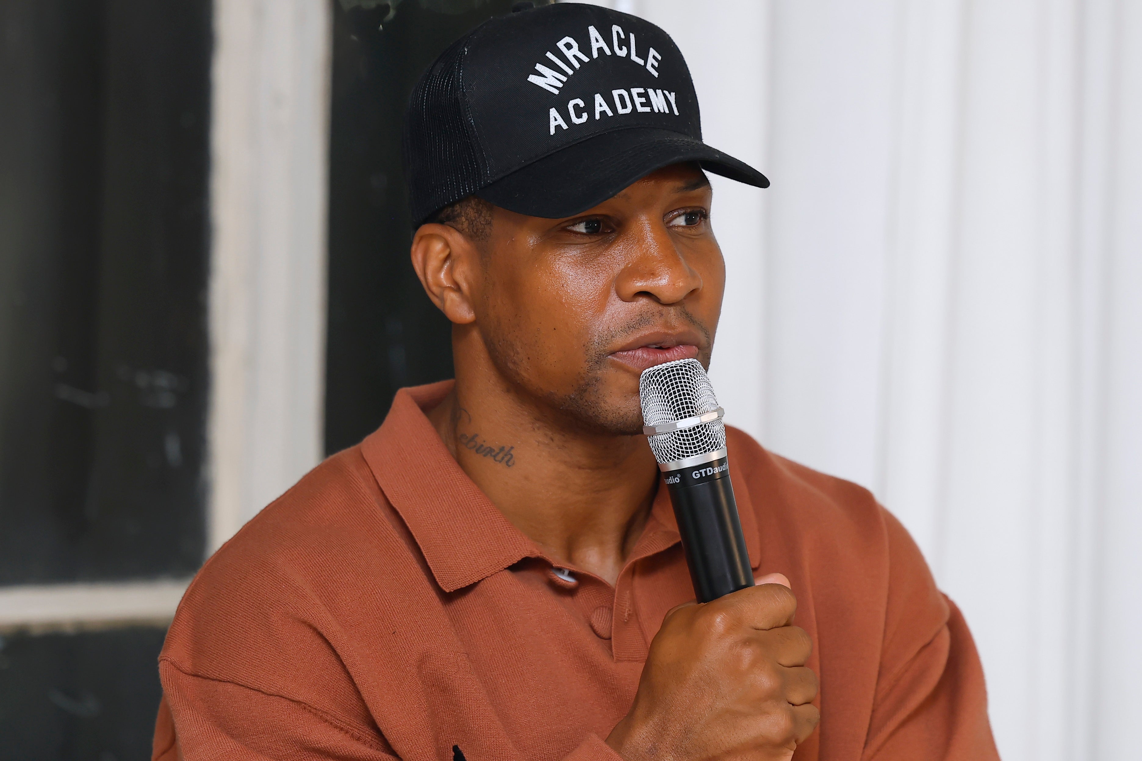 Jonathan Majors wearing a "Miracle Academy" cap, holding a microphone and speaking, dressed in casual attire